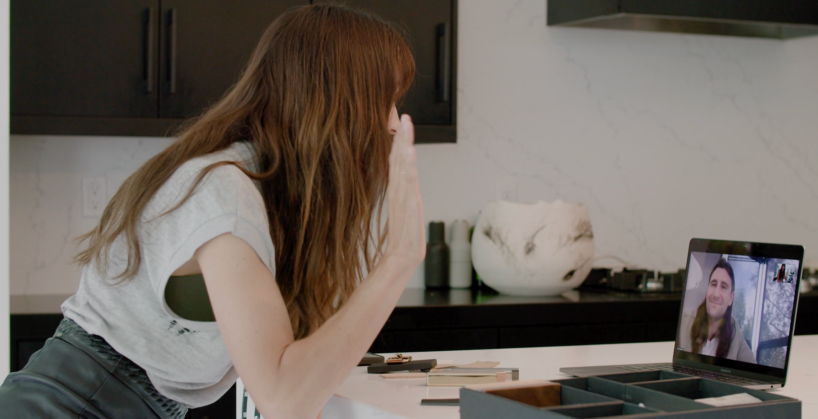 A woman waves at a laptop displaying a video call with a man.