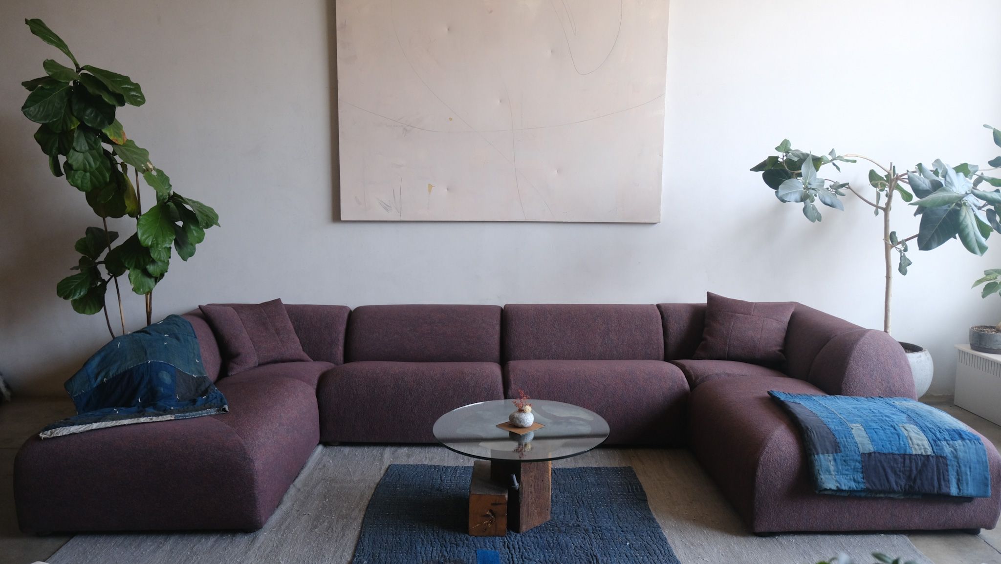 A spacious room features a plum L-shaped modular sofa with blue throws, a round glass coffee table on a blue rug, abstract wall art, and large potted plants.