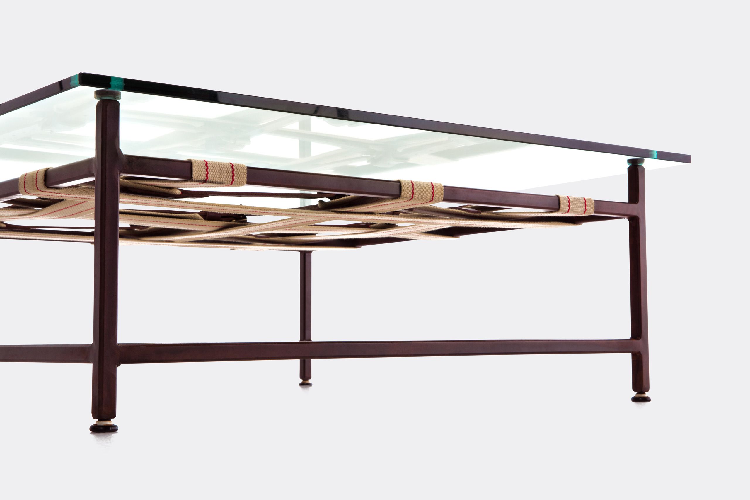 a coffee table with a glass top and a steel frame with decorative leather and webbing belts shown on a white background .