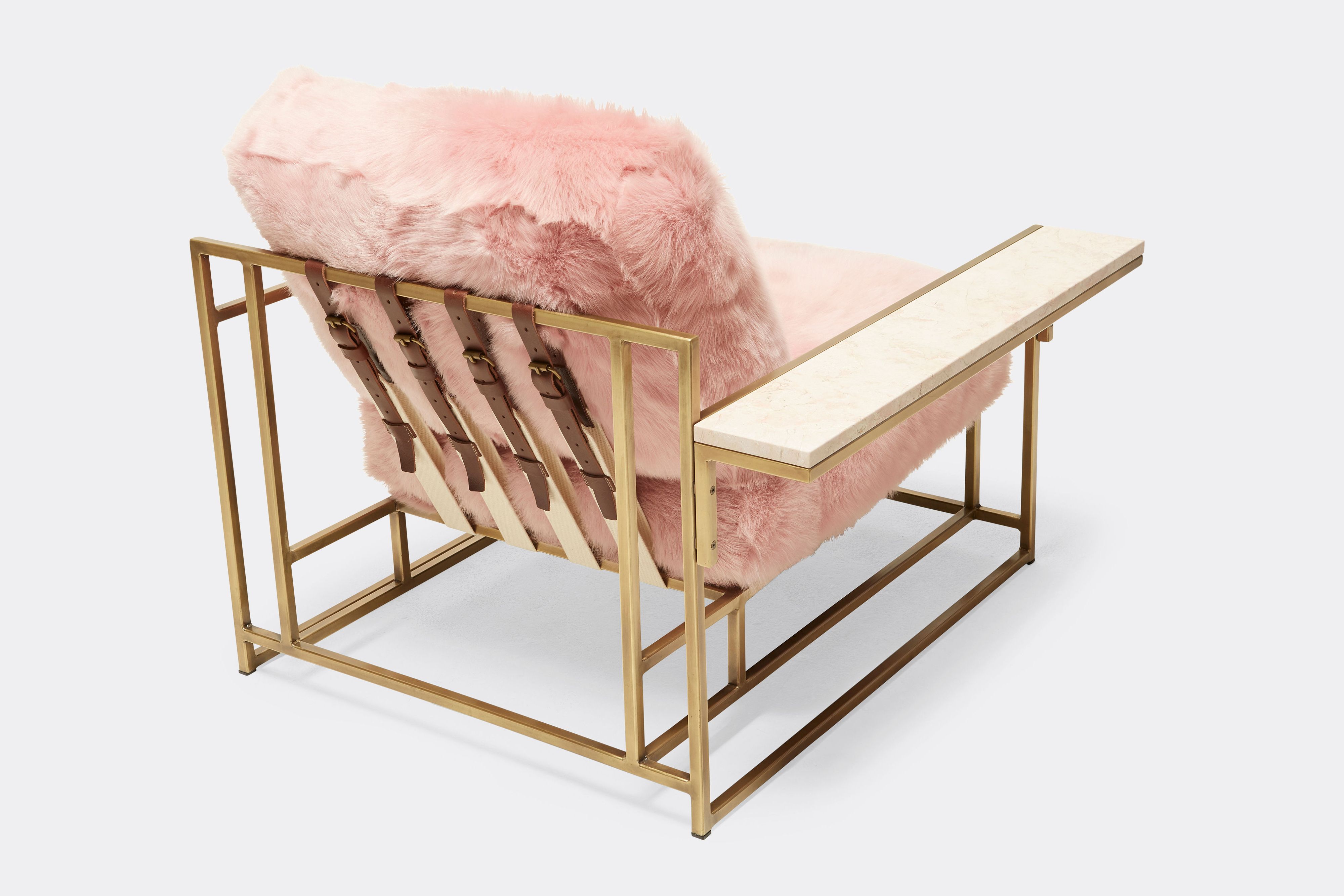 a pink shearling armchair with leather and webbing supporting belts on a brass finished steel frame and a pink marble side table attached shown on a white background .