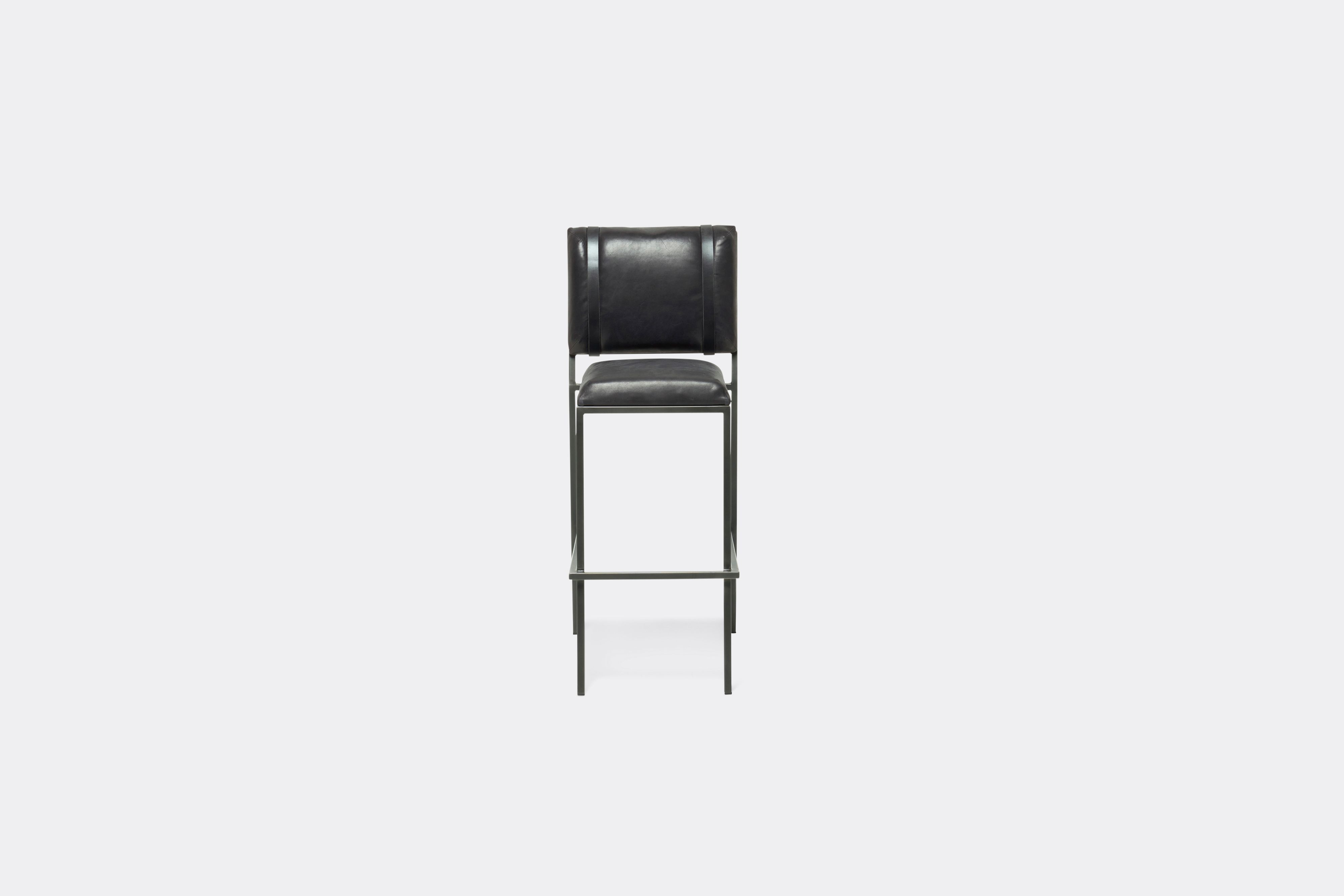 Front of barstool with black leather upholstery and black leather belts on blackened steel metal legs