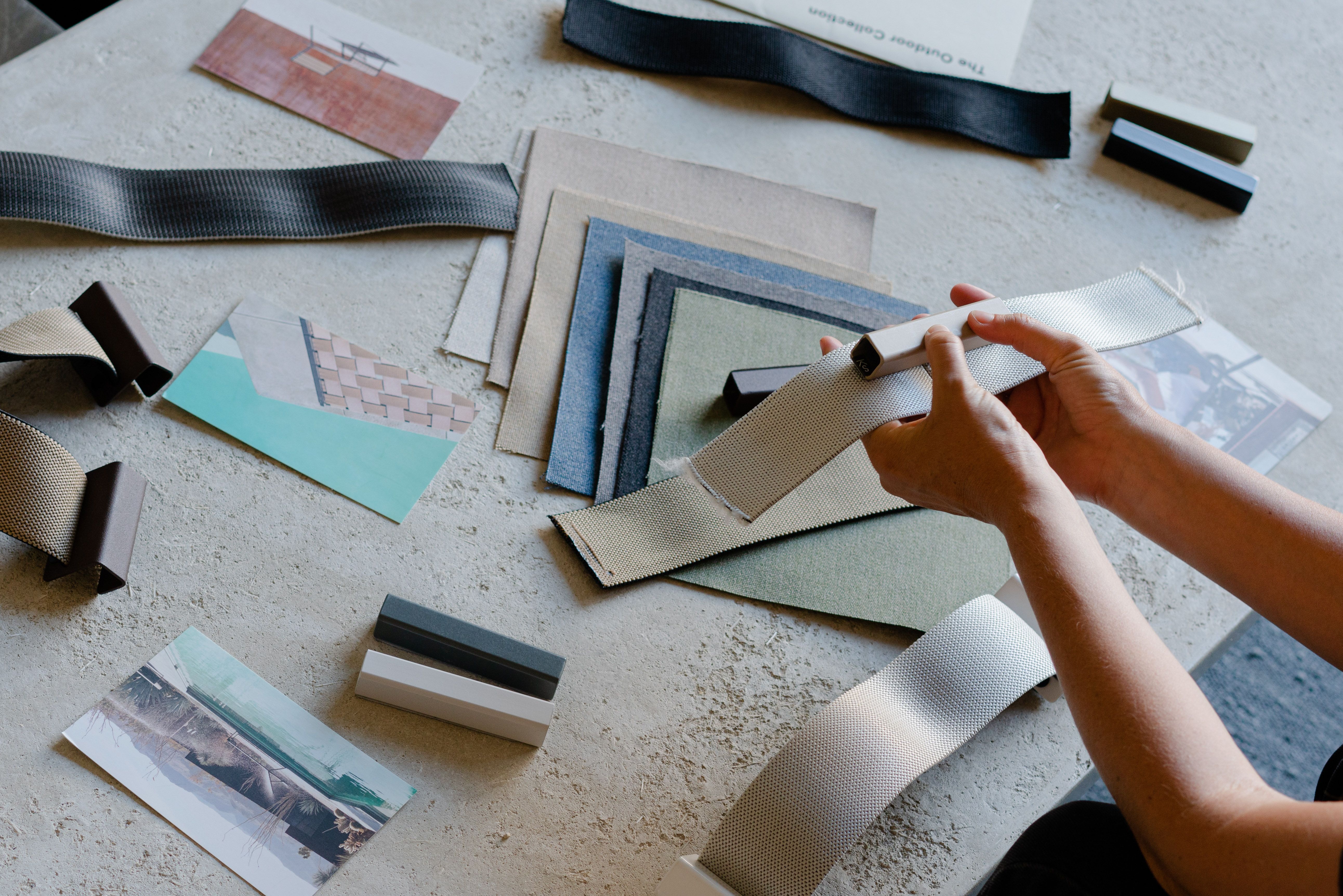 Hands holding a light gray fabric sample amidst other fabric swatches, material blocks, and design cards on a textured surface.
