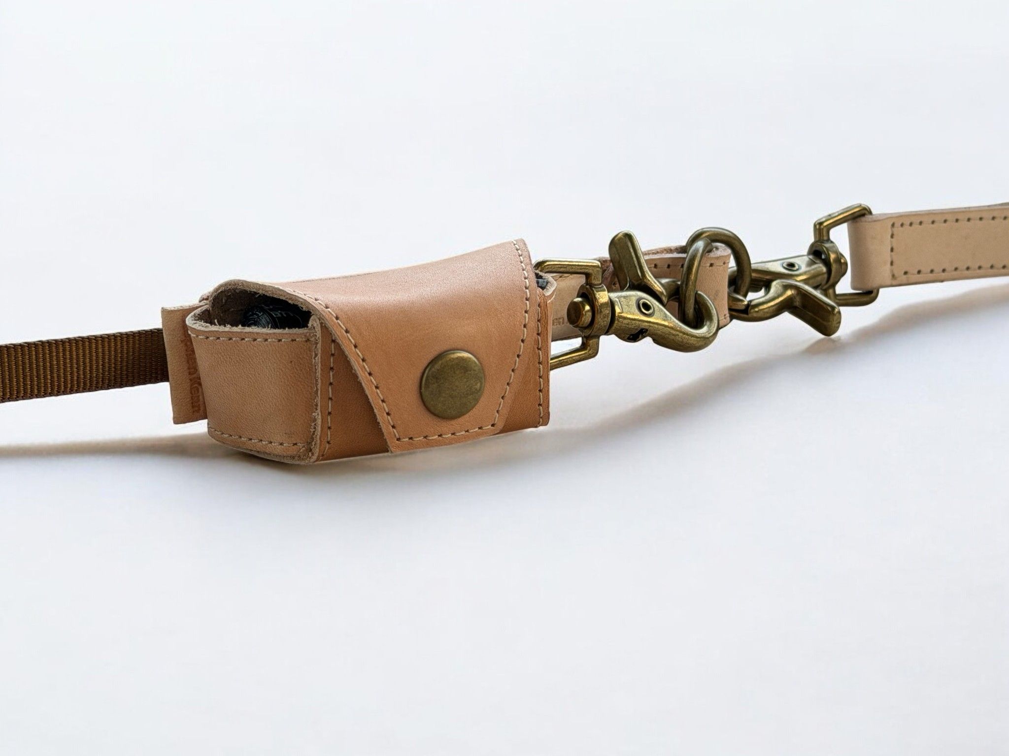 Light tan leather poop bag holder with a brass snap closure and brass clips on a strap.