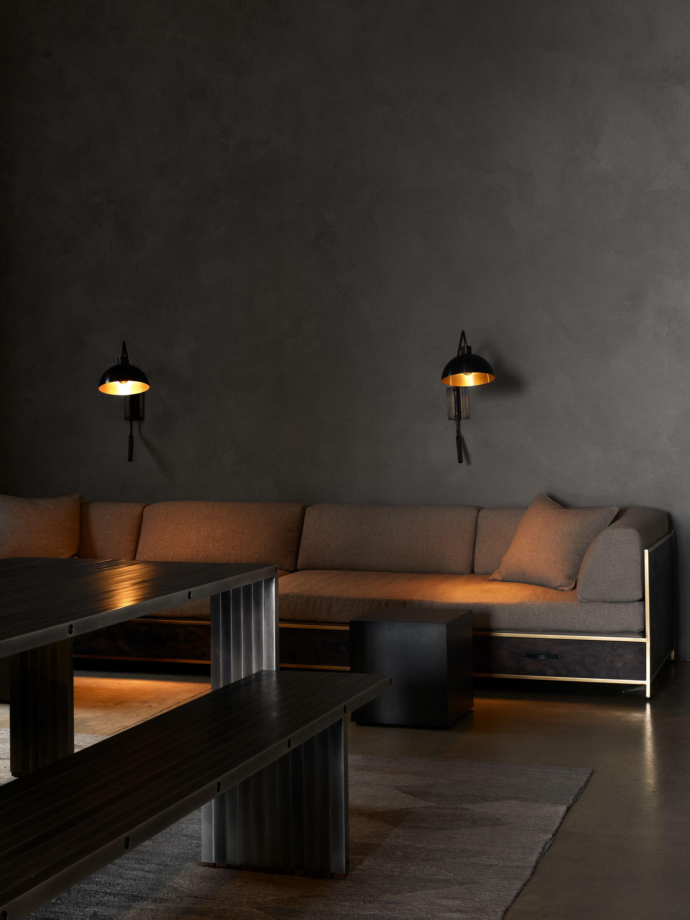 a dark room with metal dining table and a tan upholstered sofa against a grey wall with soft lights above it