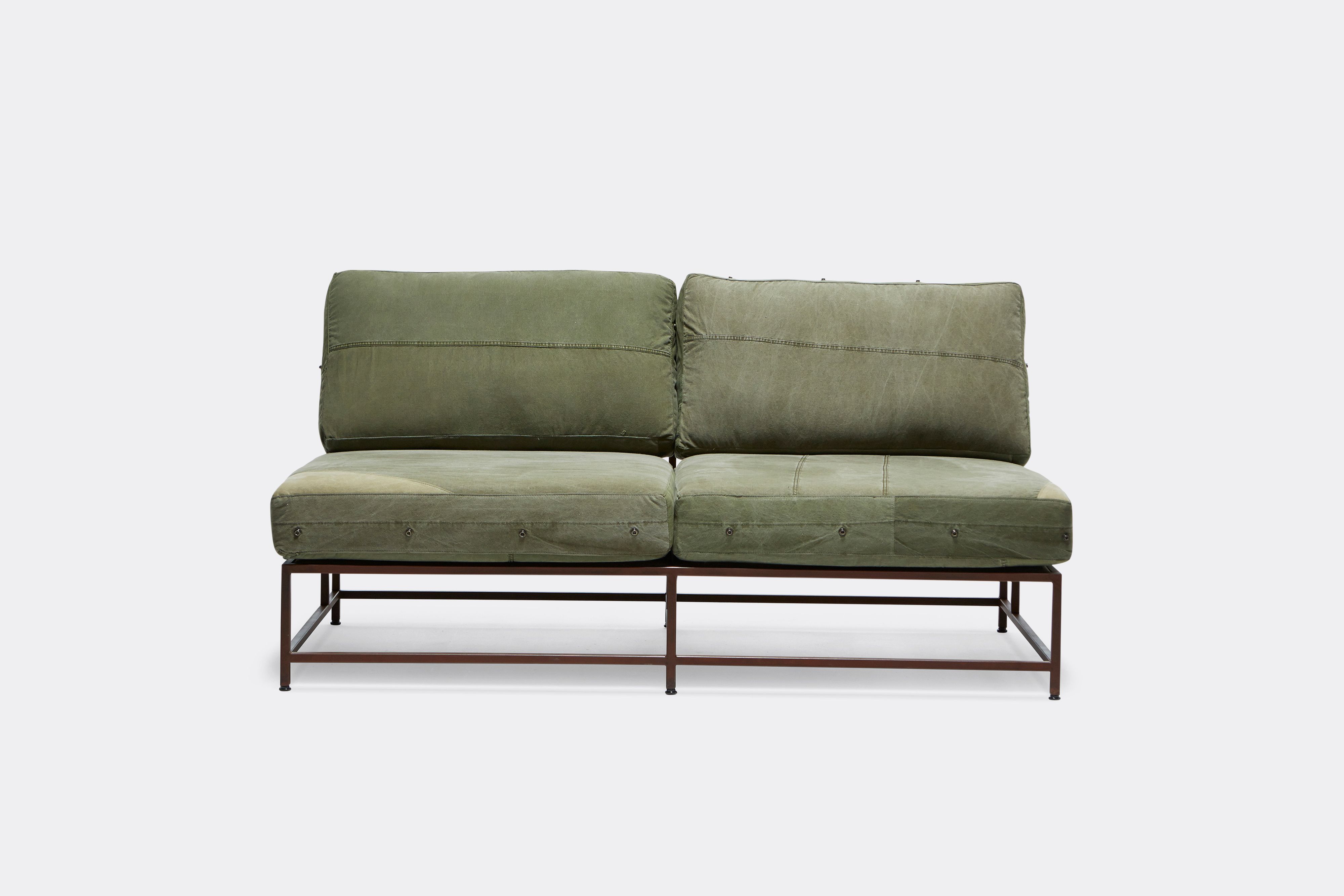 a steel framed loveseat sofa with green canvas cushions and leather and webbing supporting belts shown on a white background .