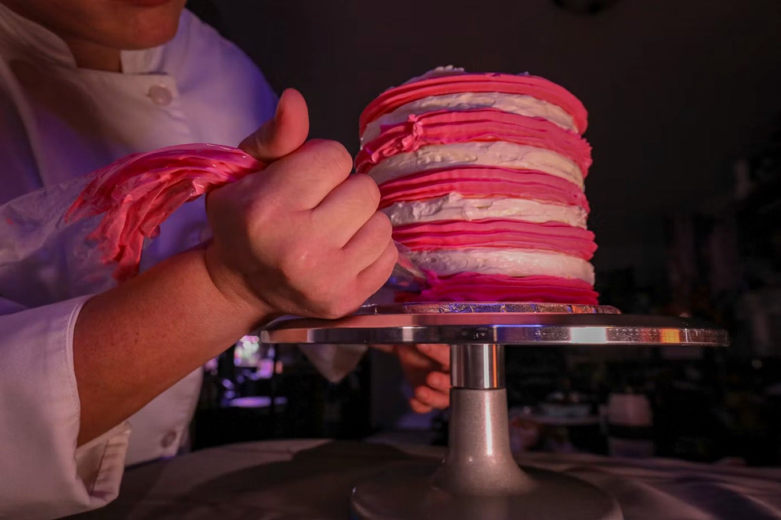 Hands piping pink buttercream onto a tall layered cake