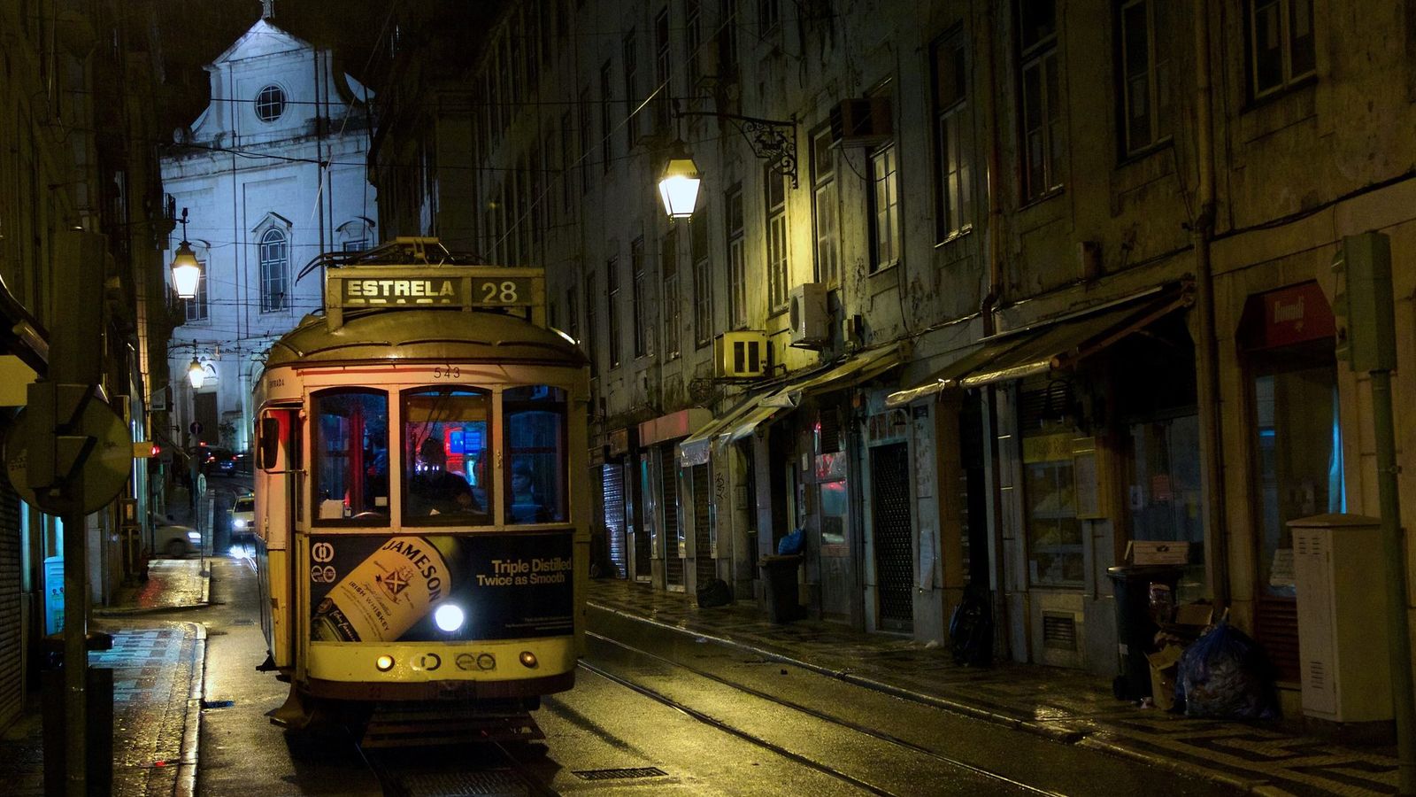 A yellow Lisbon tram on route 28 in a narrow street at night