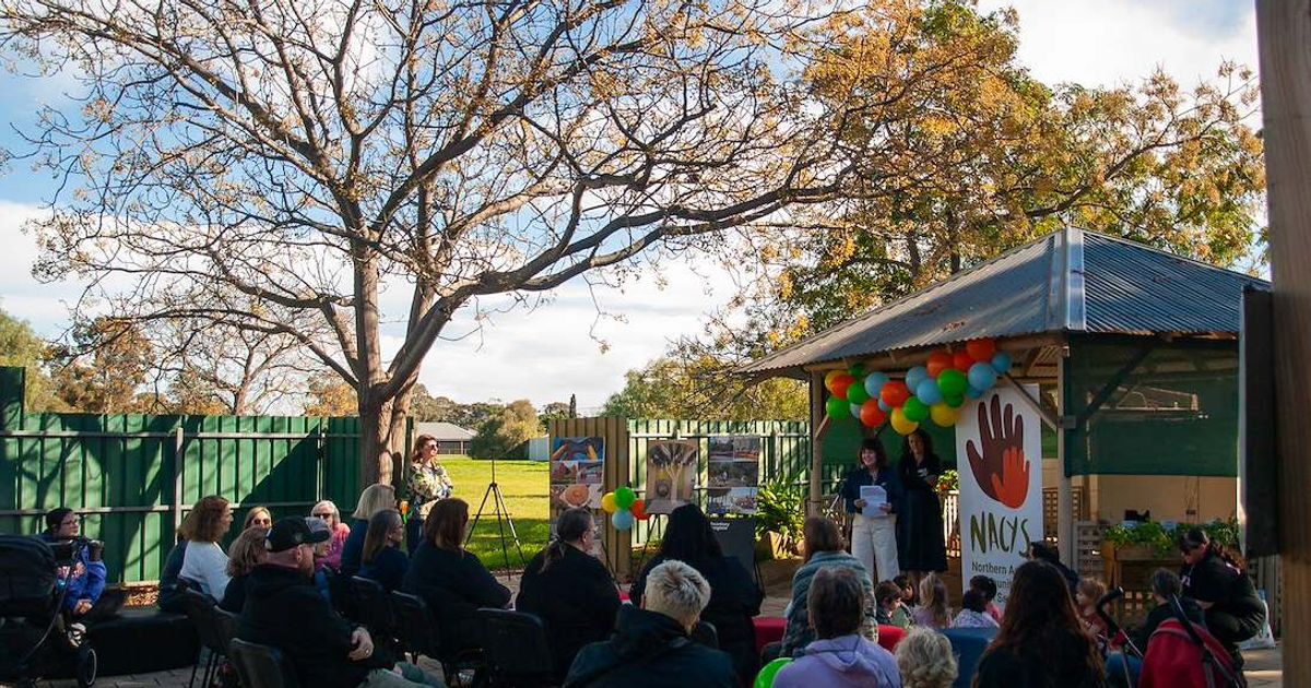 Picture of group of adults and children seated outside looking towards CEO Claire speaking at a microphone. 