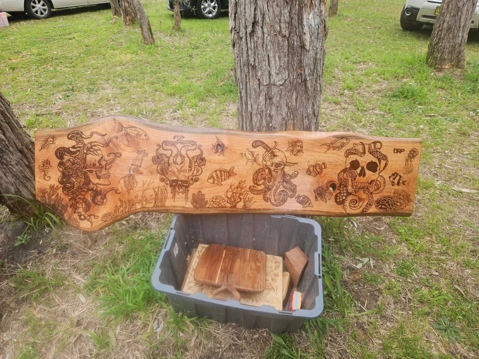 Wide natural-edged timber slab with a collection of dark character illustrations displayed outdoors