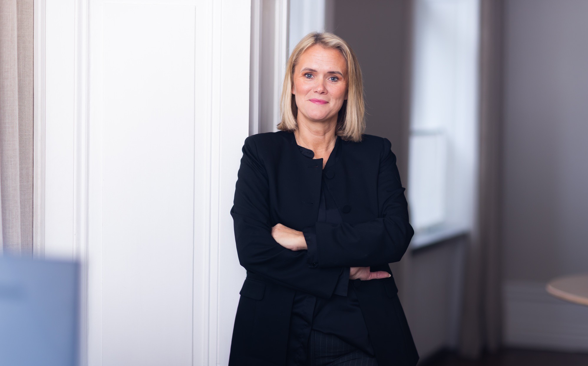 Leende kvinna i vit blus står vid höga fönster i en ljus lokal