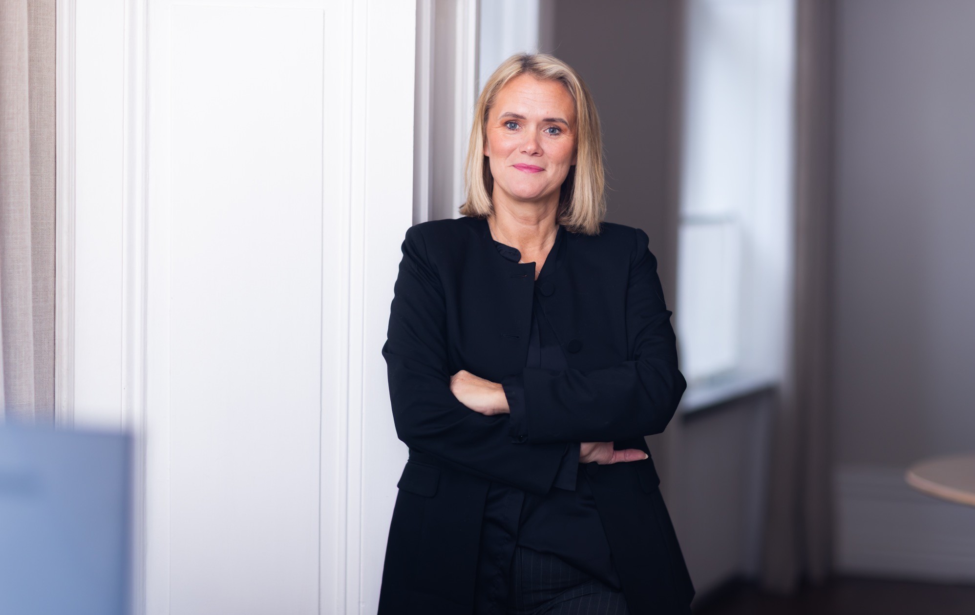 Leende kvinna i vit blus står vid höga fönster i en ljus lokal
