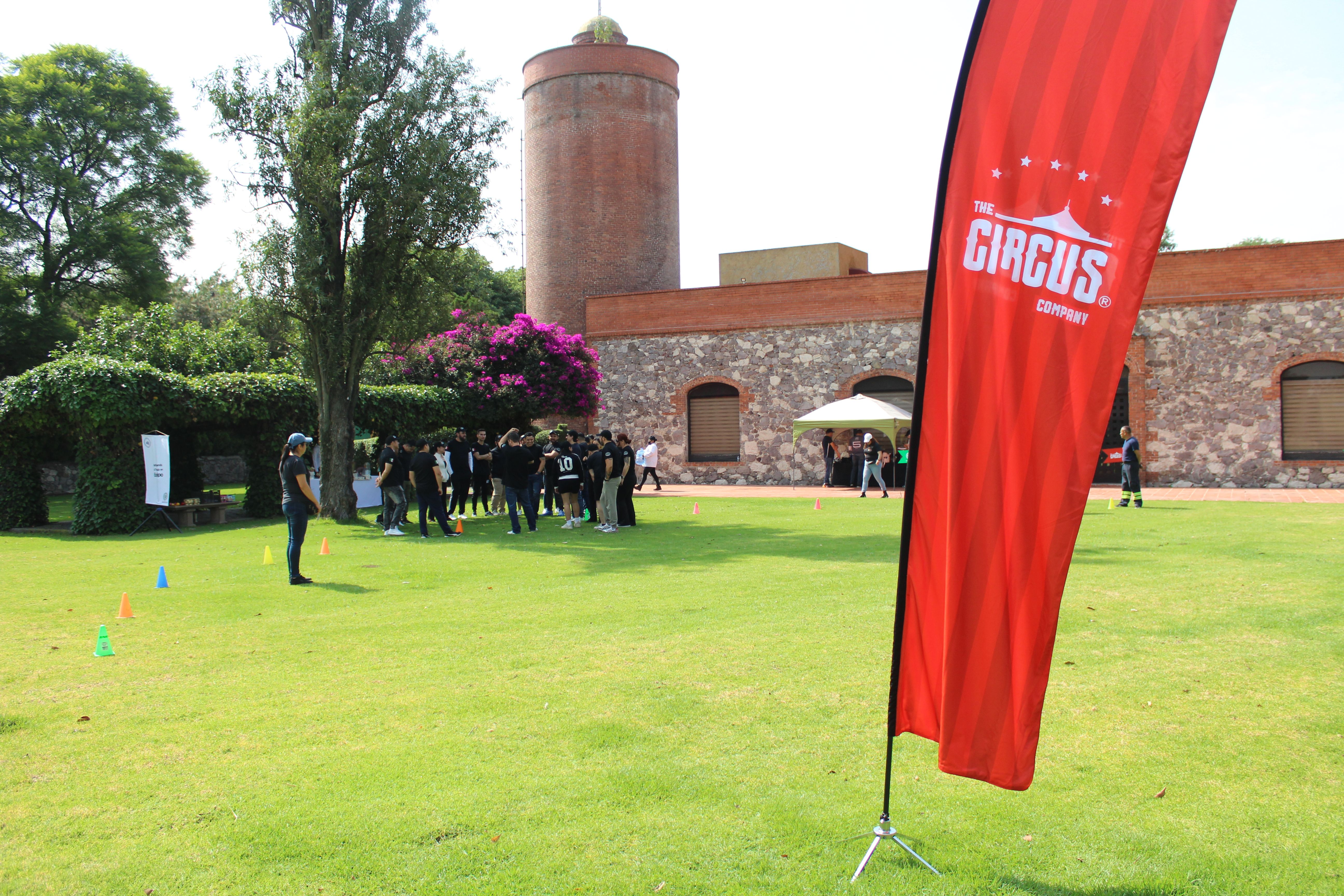 Evento al aire libre de 'The Circus Company'. Un grupo de personas reunidas en un área verde frente a un antiguo edificio de ladrillo, con una bandera roja de la marca en primer plano.