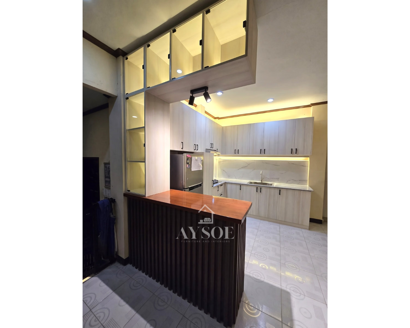 A light oak L-shaped kitchen with a marble-look backsplash, wood breakfast bar, and lit glass display cabinets.