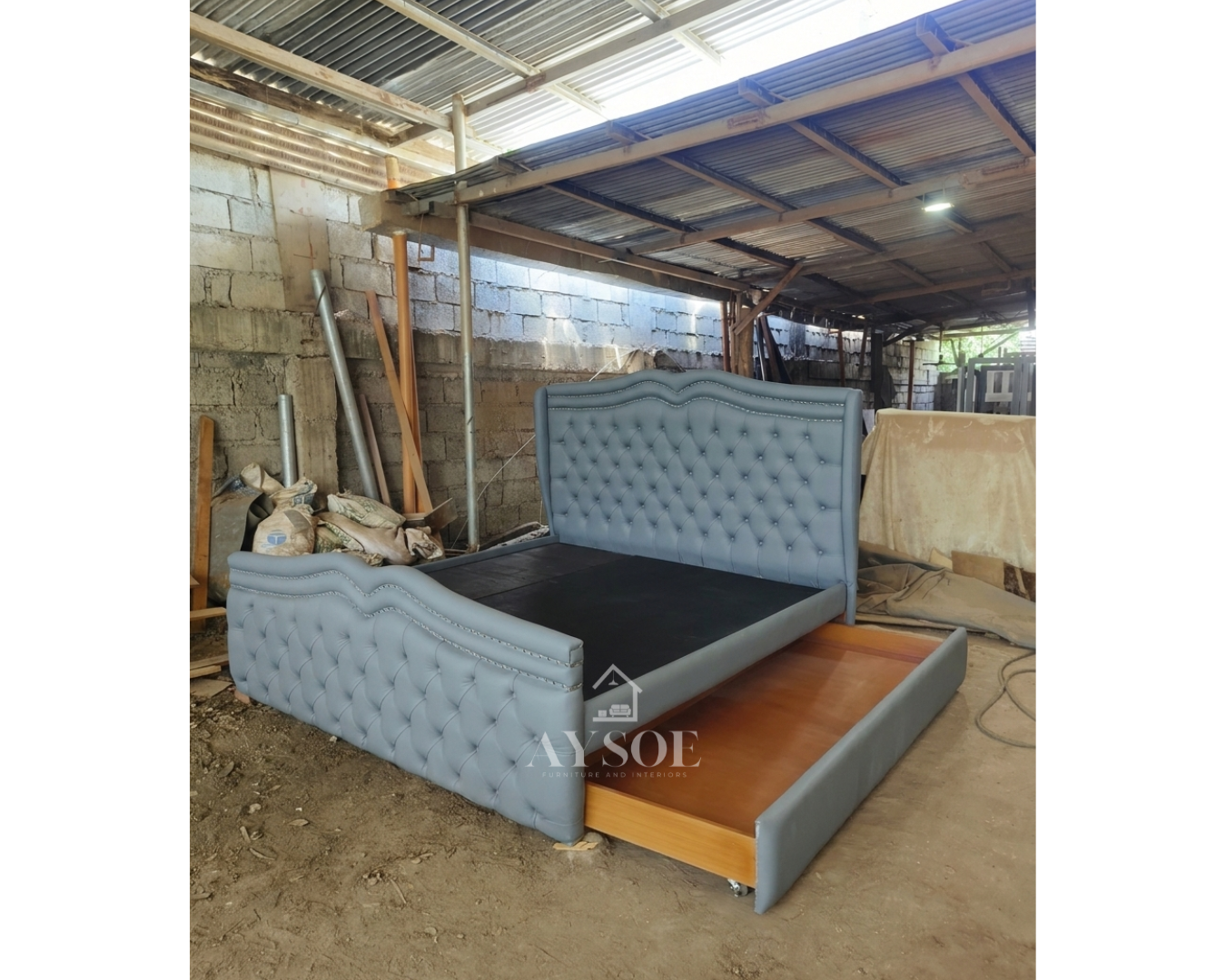 A luxurious bed frame in slate blue-grey leatherette, with a tall tufted wingback headboard, a matching footboard, nailhead trim, and a roll-out trundle drawer. Elegant and space-saving.