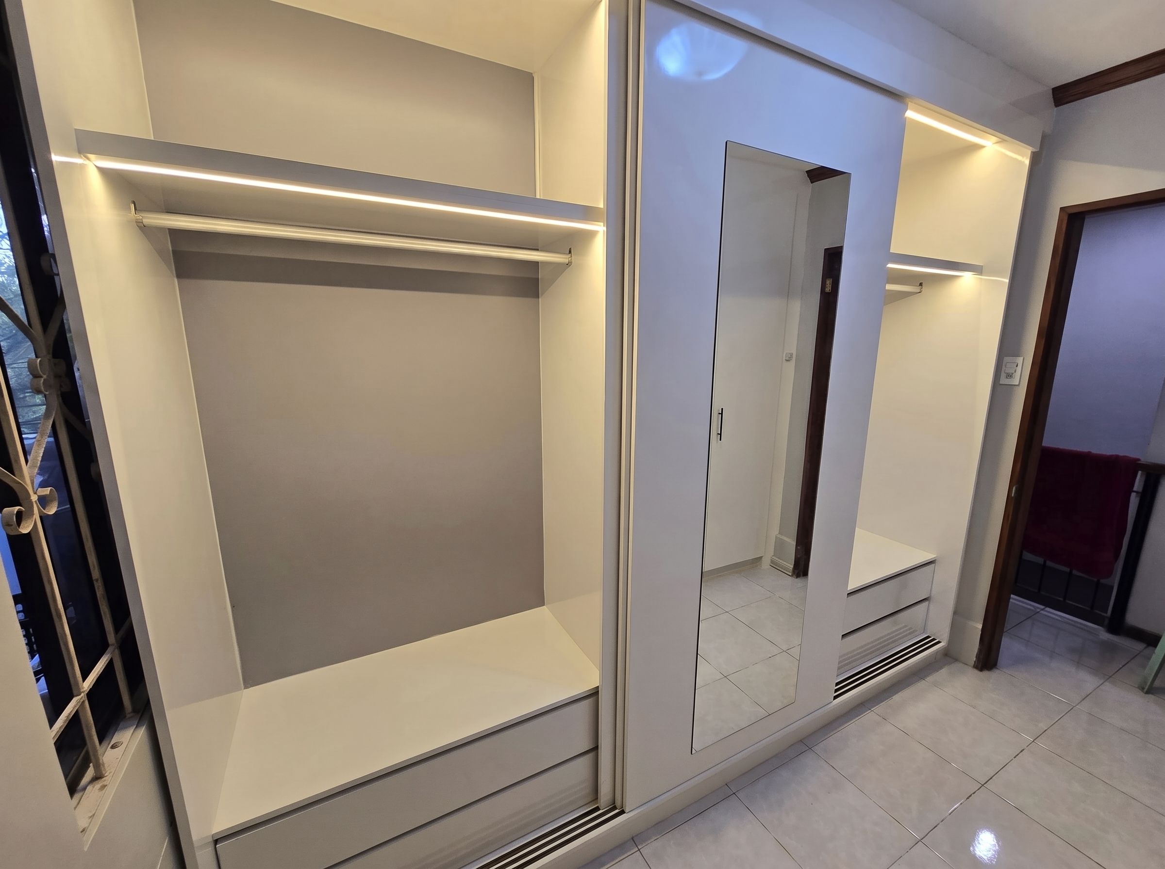 A white built-in wardrobe with a sliding mirror-panel door, two wide hanging sections, four deep drawers, and warm LED lighting inside. Made to fit your wall.
