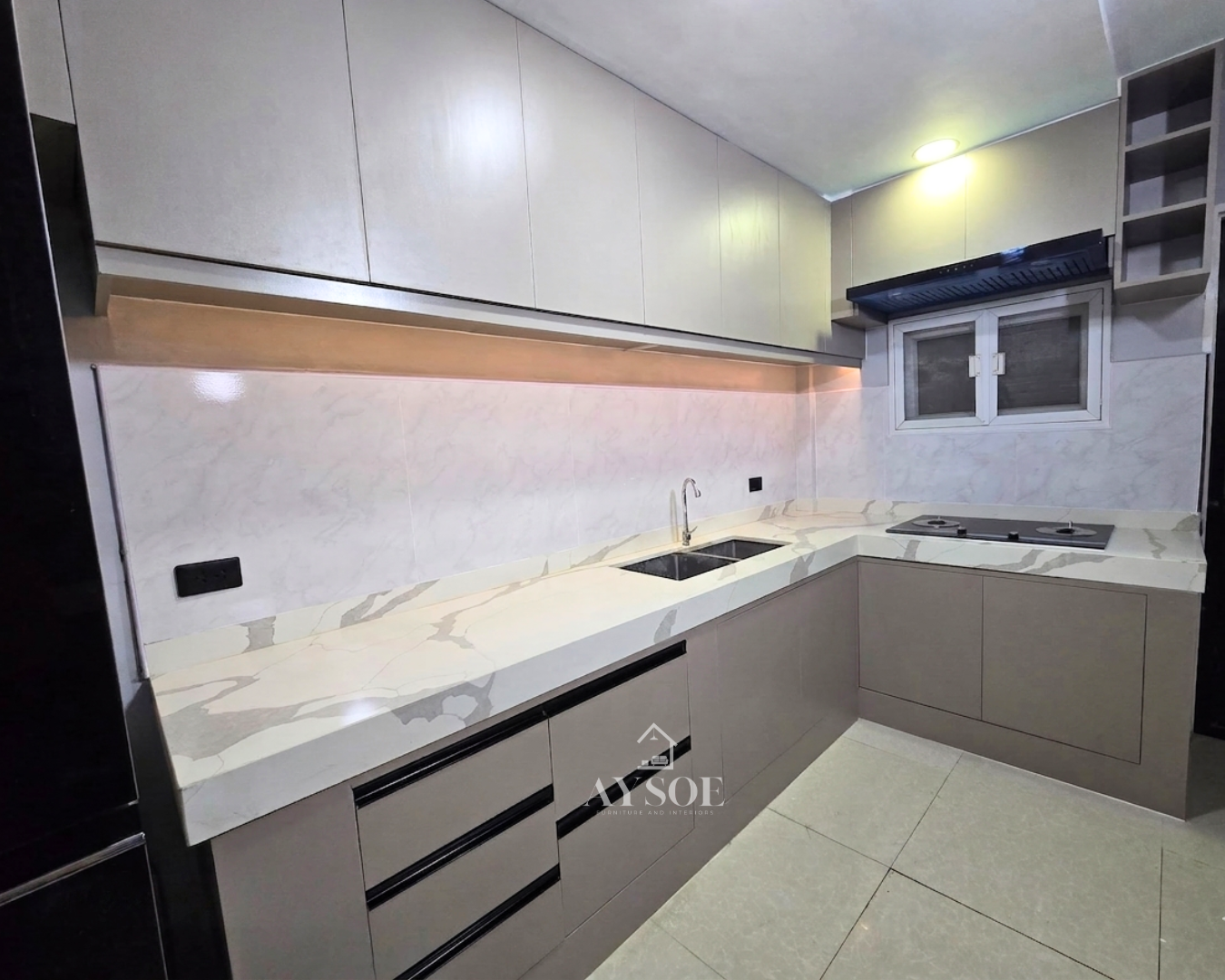 A modern L-shaped kitchen set with cream upper cabinets, taupe lower cabinets, and a marble-look countertop. Handleless, bright, and full of storage.