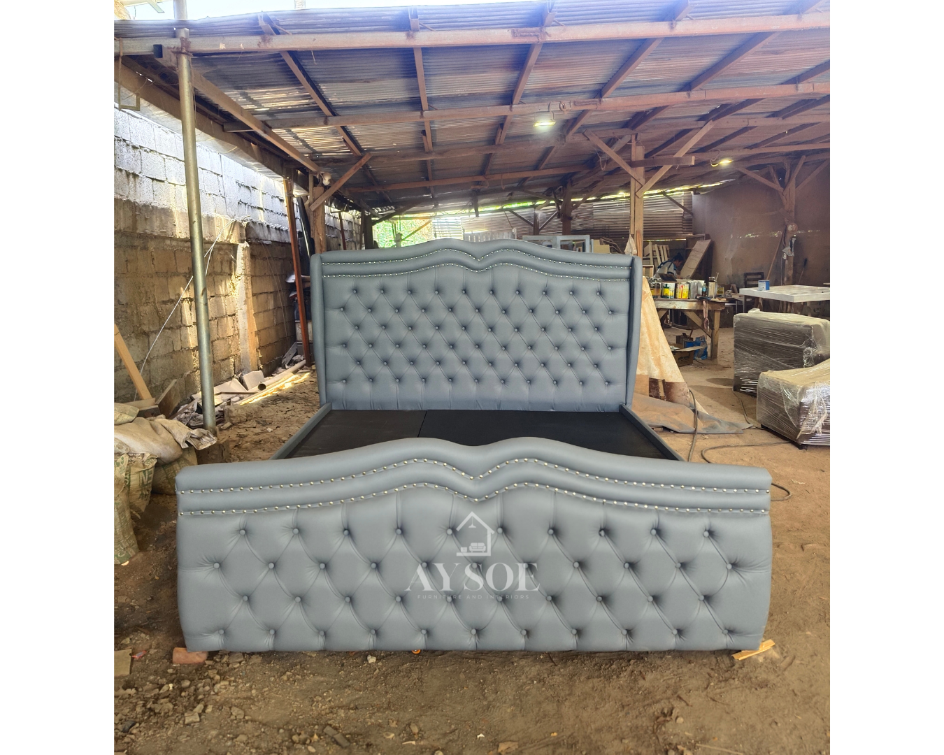 A luxurious bed frame in slate blue-grey leatherette, with a tall tufted wingback headboard, a matching footboard, nailhead trim, and a roll-out trundle drawer. Elegant and space-saving.