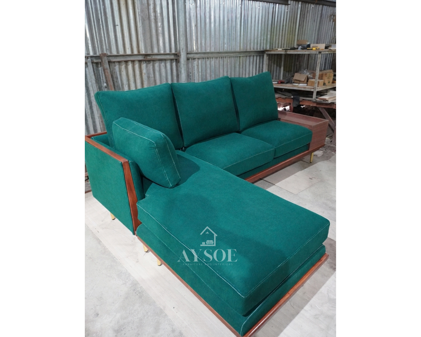 A mid-century L-shaped sofa in rich emerald velvet-style fabric, with a deep left chaise, plush loose cushions, a walnut base, brass-gold legs, and a built-in storage side table. Bold, comfortable, made to order.