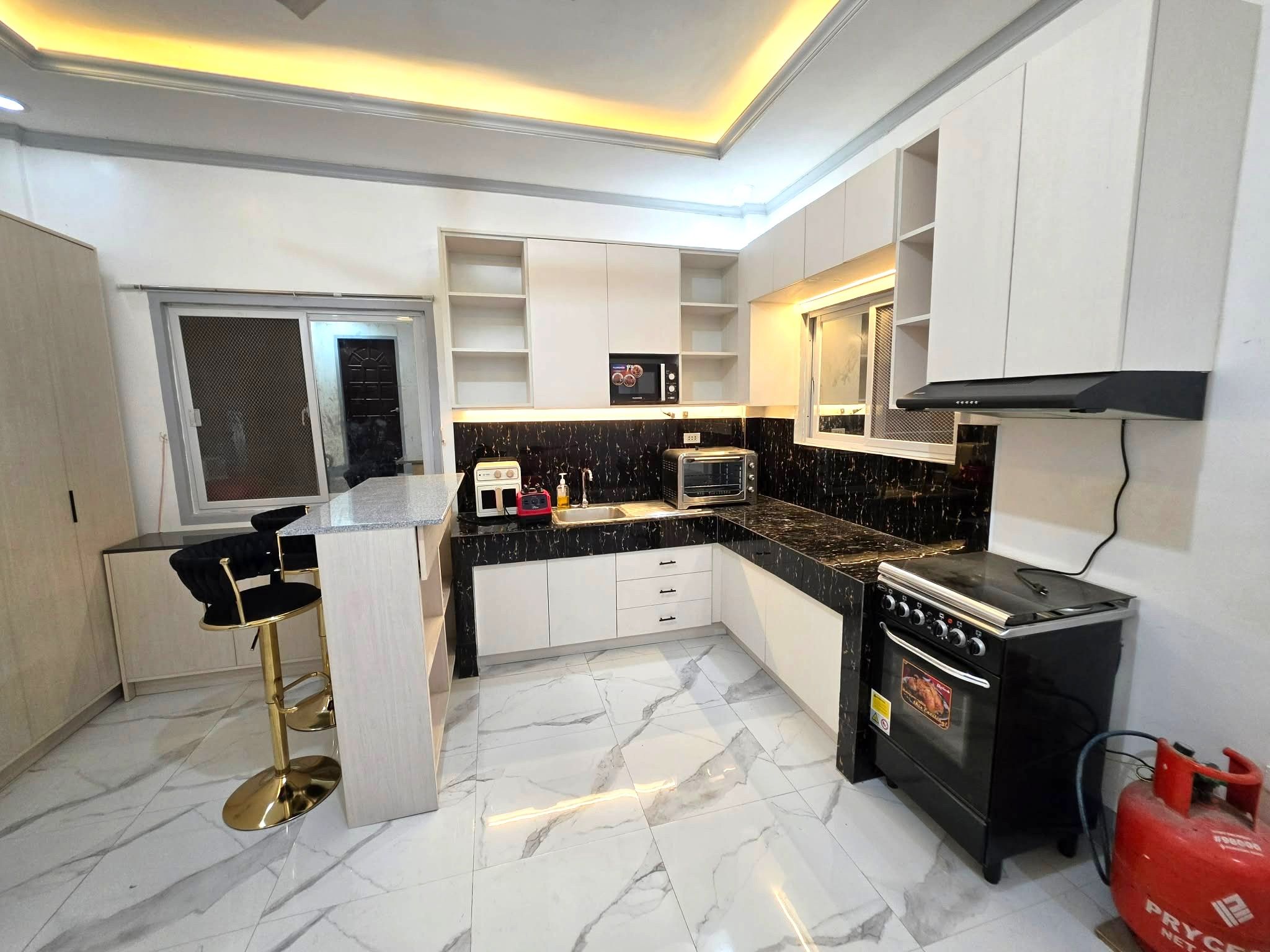 A cream wood-grain L-shaped kitchen with a black and gold backsplash, open shelves, and a granite-top breakfast bar.