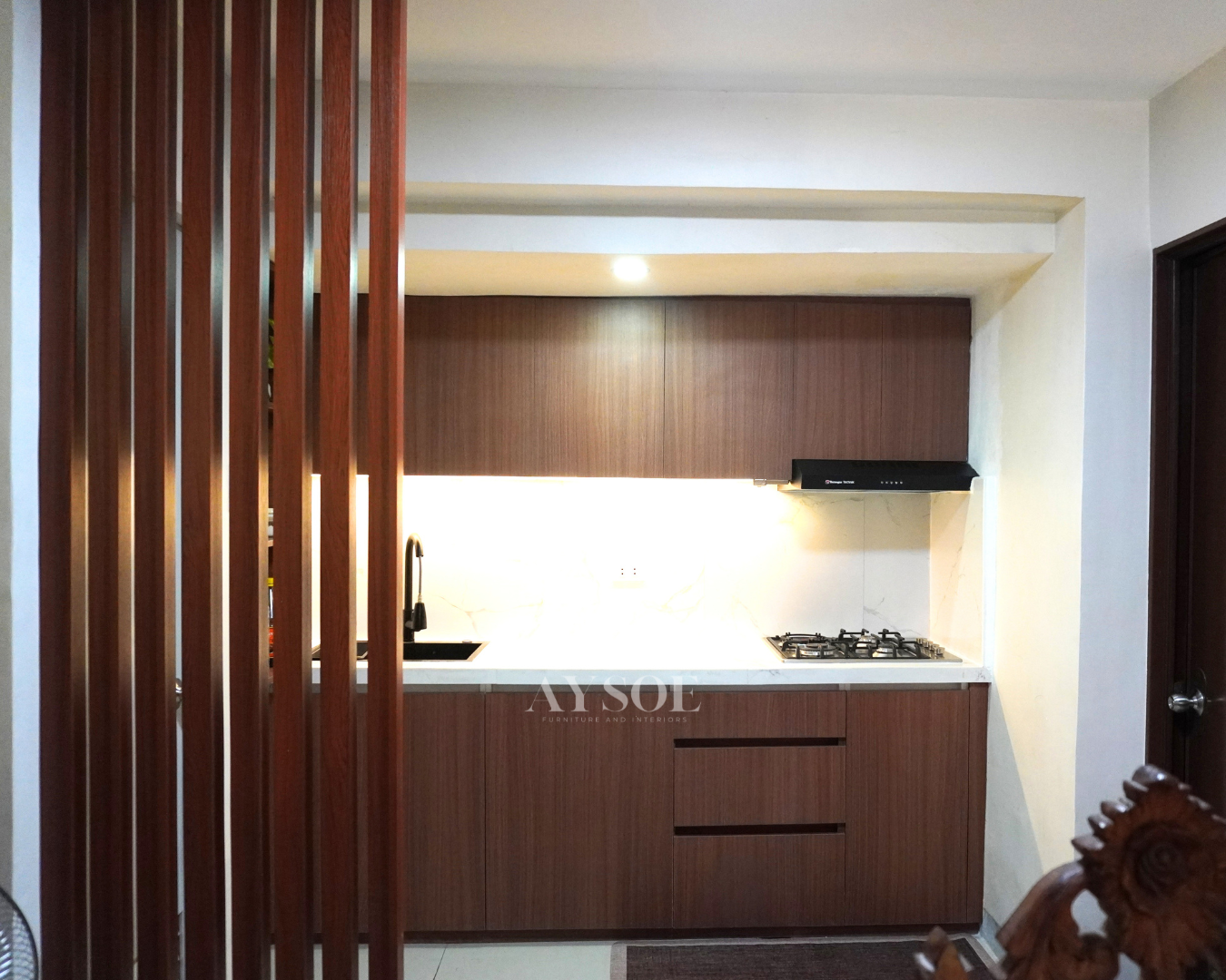 A single-wall kitchen in rich walnut-grain cabinets with a marble-look backsplash. Handleless doors, a black matte sink, and under-cabinet lighting make it cozy and modern.