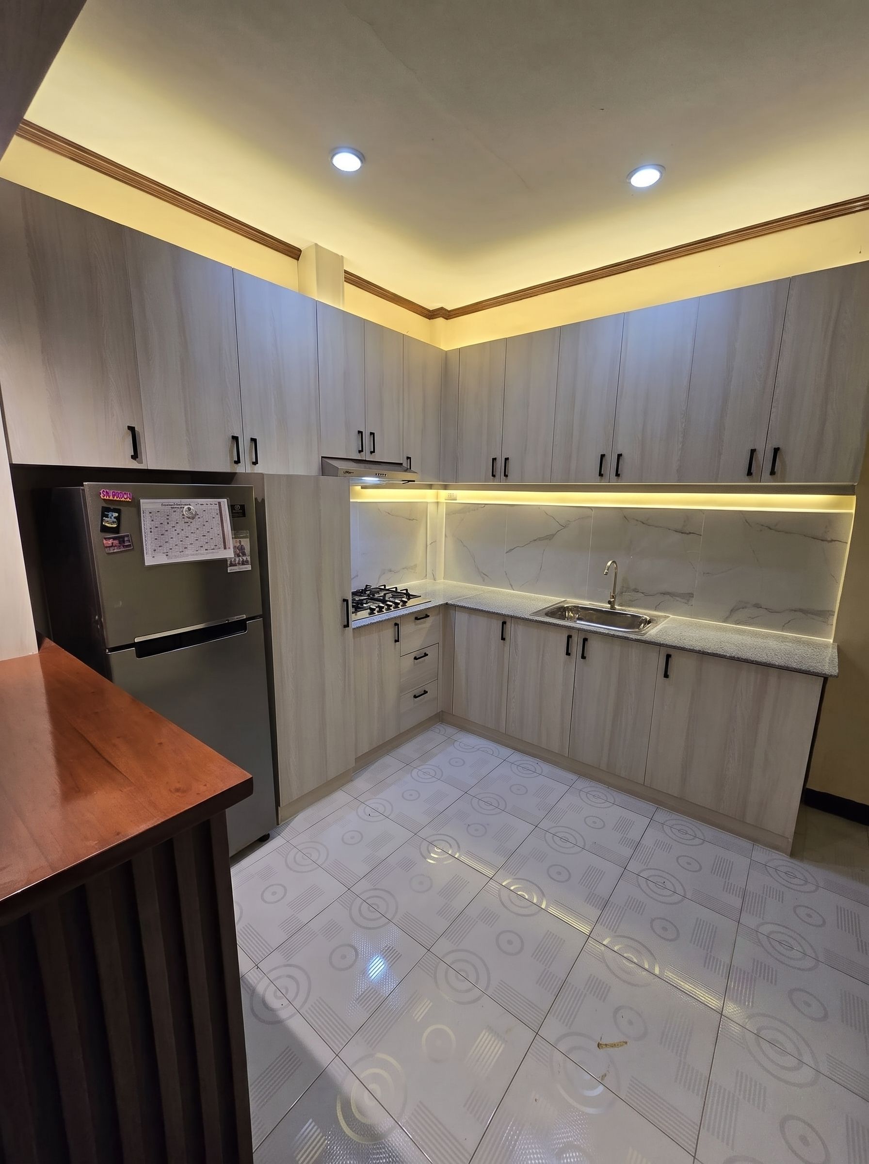 A light oak L-shaped kitchen with a marble-look backsplash, wood breakfast bar, and lit glass display cabinets.