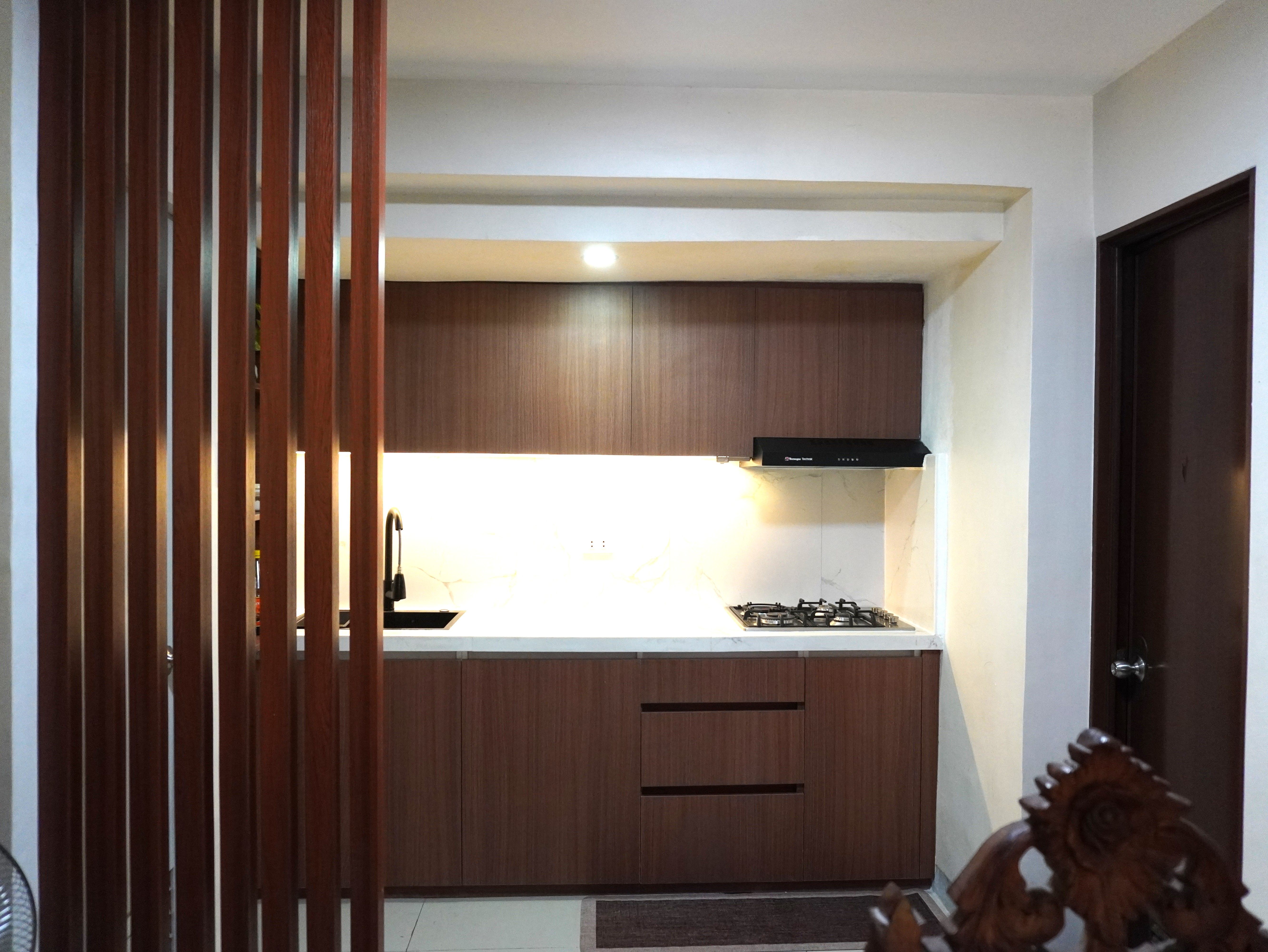 A warm single-wall kitchen in rich walnut-grain cabinetry with a Calacatta-style marble backsplash. Handleless doors, a black matte sink and faucet, and glowing under-cabinet lighting make it feel cozy and modern.