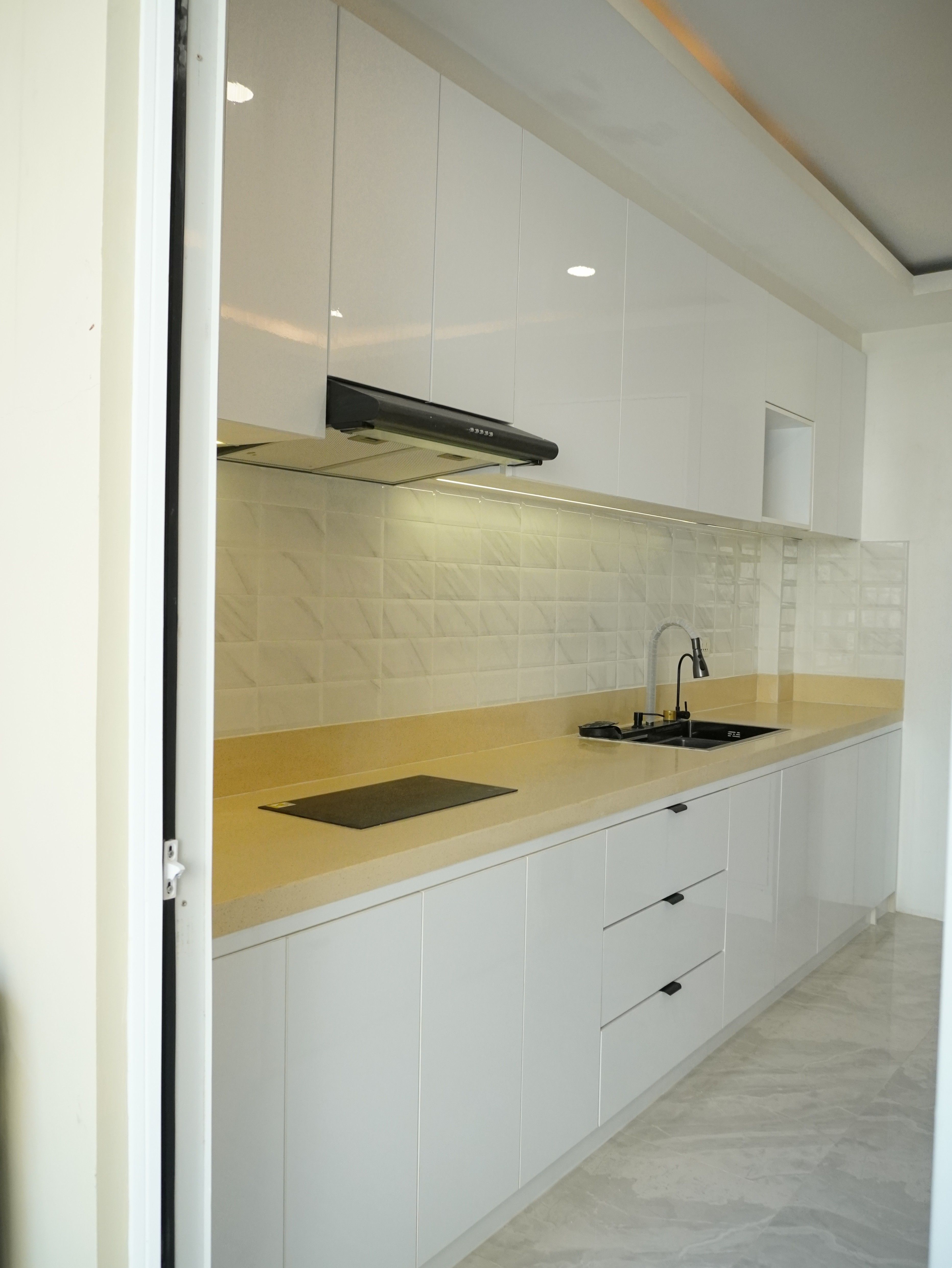 A full-room kitchen in high-gloss white with a waterfall island, sand-toned solid-surface counters, and a cove-lit tray ceiling. Floor-to-ceiling cabinetry, a built-in induction hob, and a tall pantry unit keep everything organized in a bright, open layout.