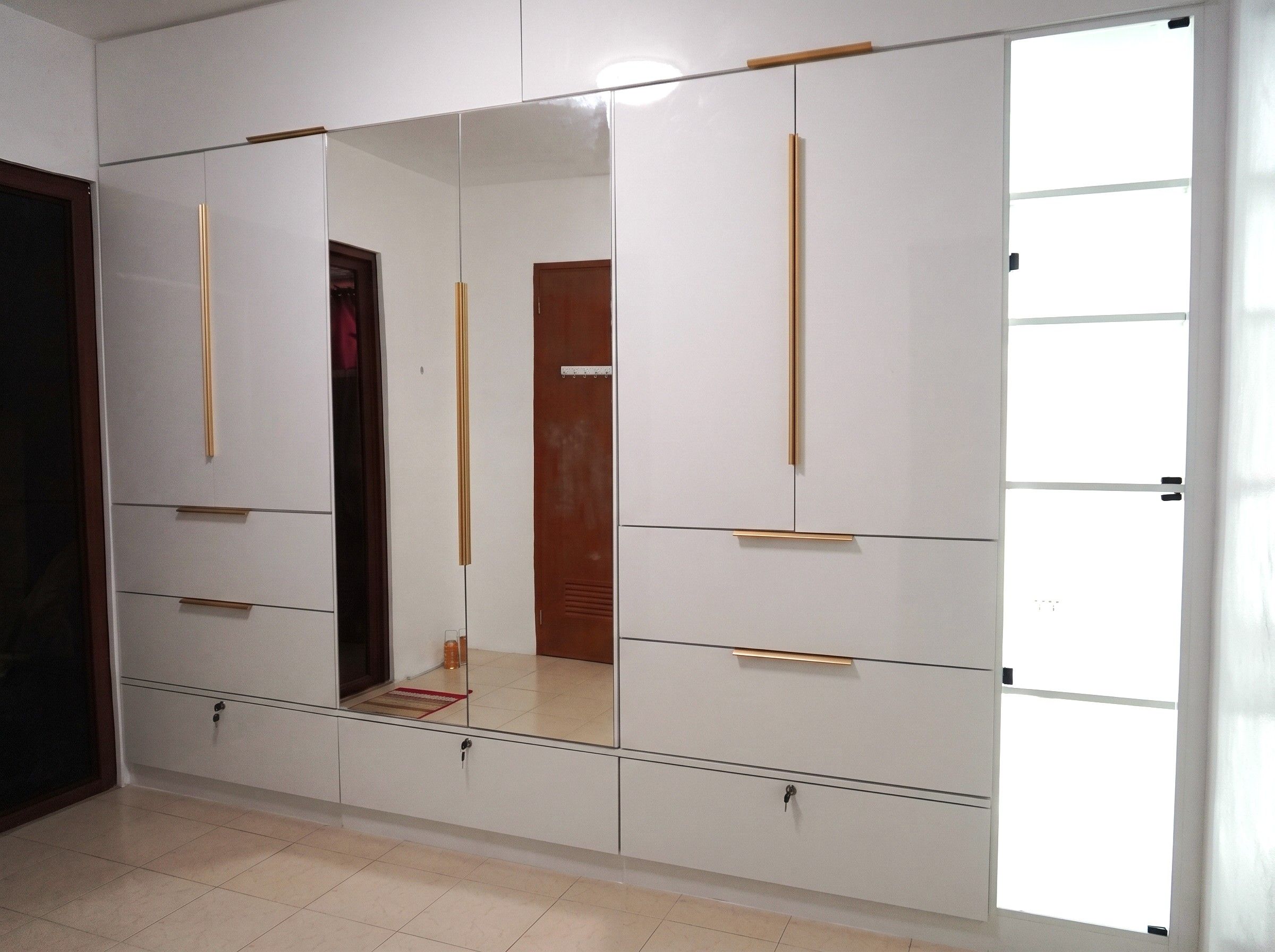 A floor-to-ceiling built-in wardrobe in high-gloss white, with gold bar handles, a full-height center mirror, roomy drawers, three locking drawers, and glass-door shelving. Made to fit your wall.