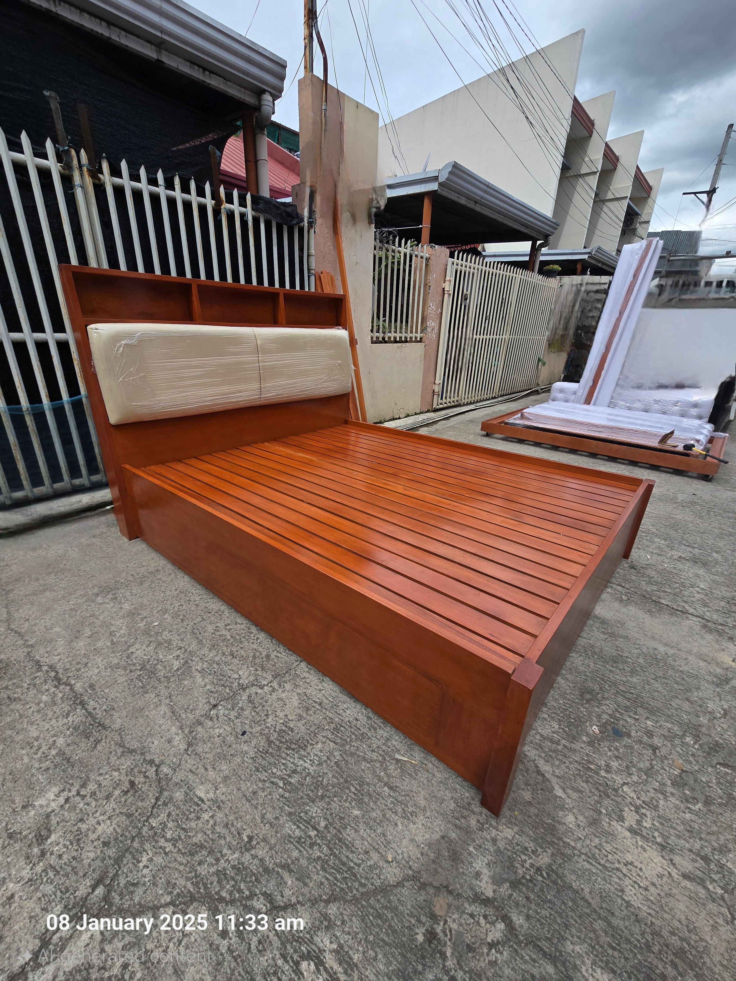 A solid wood bed in a rich cherry finish, with a built-in shelf headboard for books, lamps, and a phone, and cream padded panels for comfortable back support. Clean lines, warm tones, and a sturdy frame.