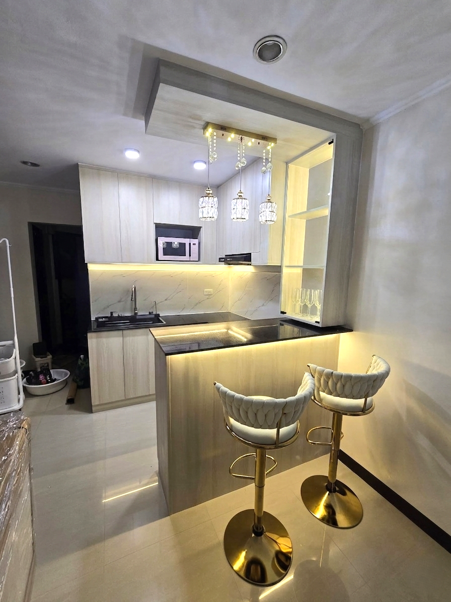 A compact light oak kitchen with a marble-look backsplash, breakfast bar, and lit glass display cabinet.