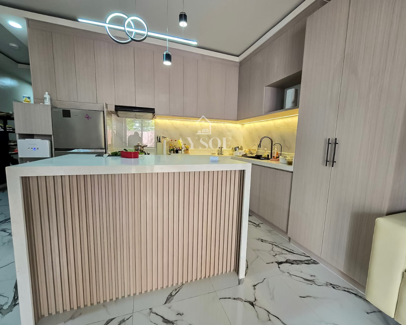 An L-shaped kitchen in soft taupe wood-grain cabinets with a marble-look backsplash and warm LED lighting. A white waterfall island with a wood-slat front and tall storage complete the look.
