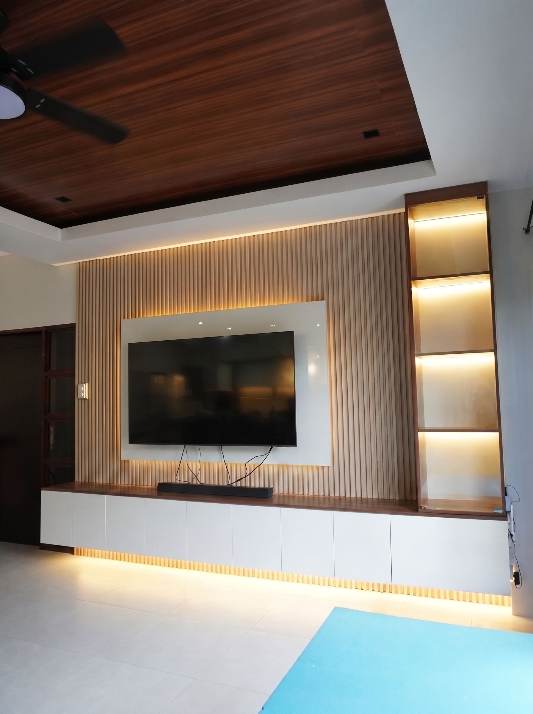 A built-in TV wall with a vertical wood-slat feature panel, a white gloss TV backing, a long floating storage cabinet, and a tall glass-door display tower. Warm LED lighting makes it glow. Made to fit your wall.