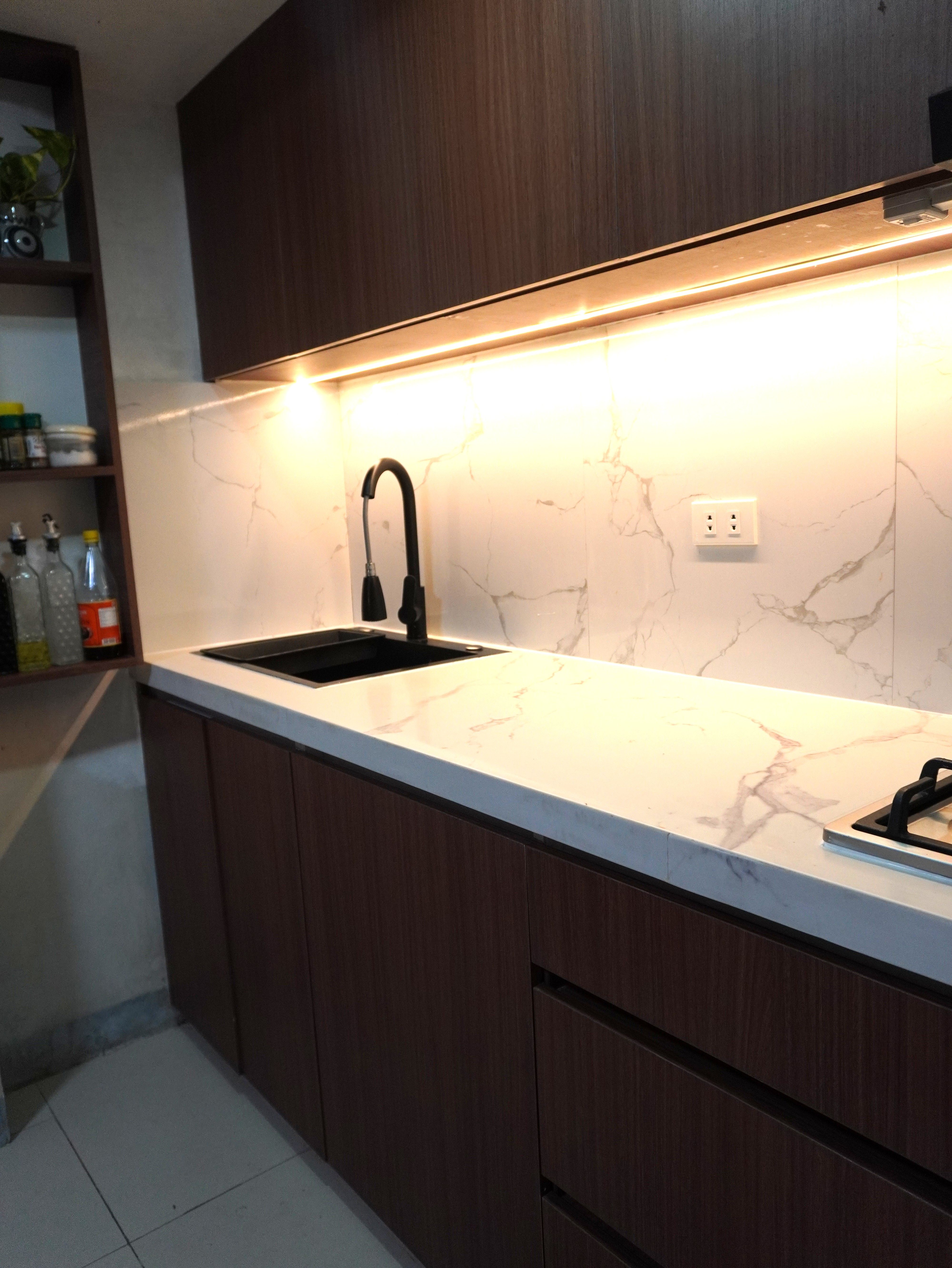 A warm single-wall kitchen in rich walnut-grain cabinetry with a Calacatta-style marble backsplash. Handleless doors, a black matte sink and faucet, and glowing under-cabinet lighting make it feel cozy and modern.