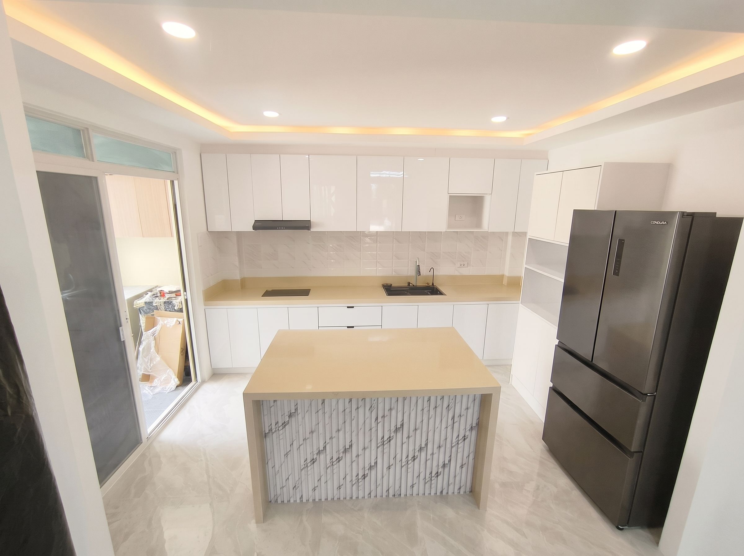 A full-room kitchen in high-gloss white with a waterfall island, sand-toned solid-surface counters, and a cove-lit tray ceiling. Floor-to-ceiling cabinetry, a built-in induction hob, and a tall pantry unit keep everything organized in a bright, open layout.