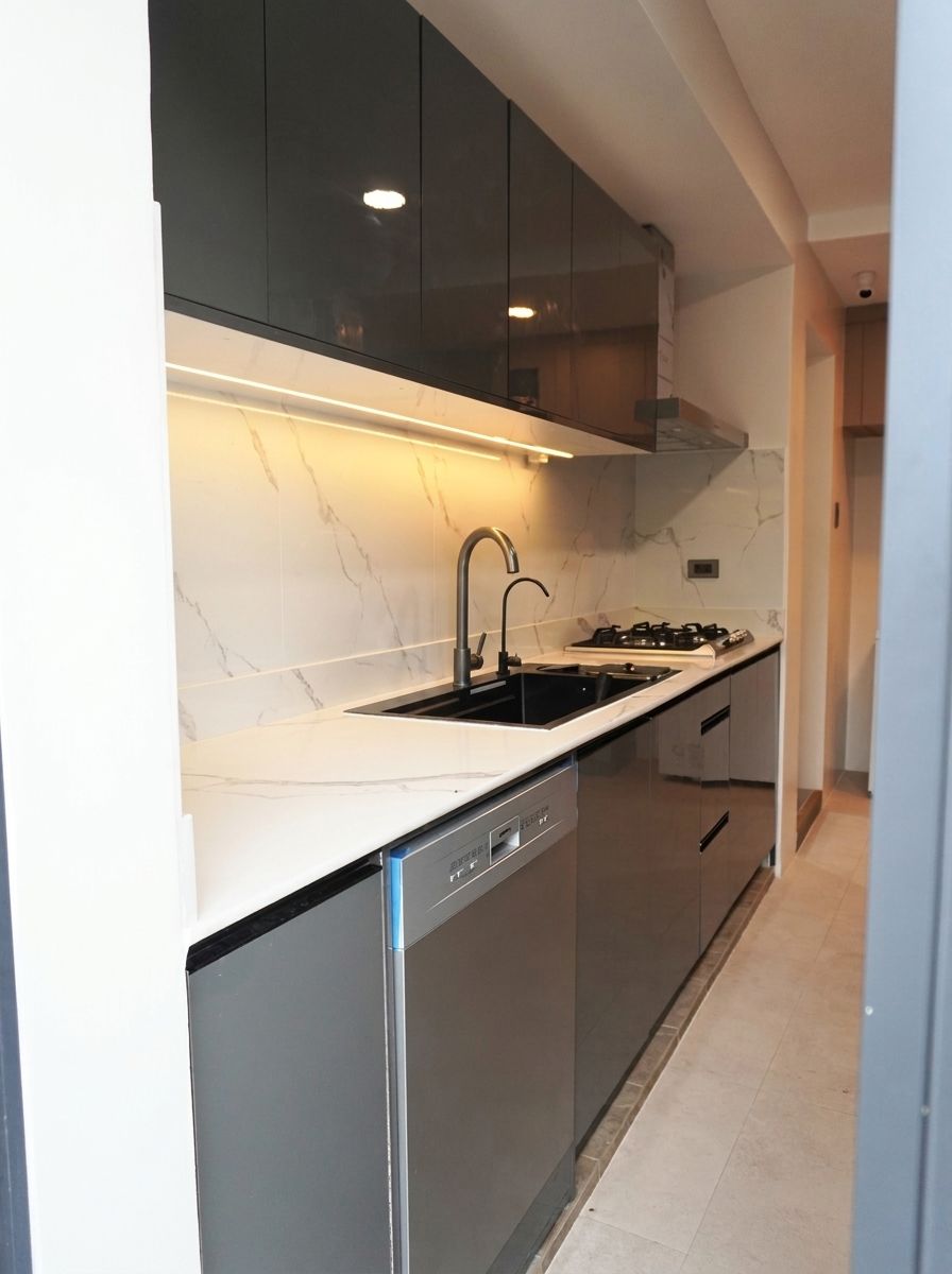 A sleek single-wall kitchen in high-gloss charcoal with a Calacatta-style marble backsplash. Handleless cabinetry, integrated appliances, and warm under-cabinet lighting give it a clean, modern look.