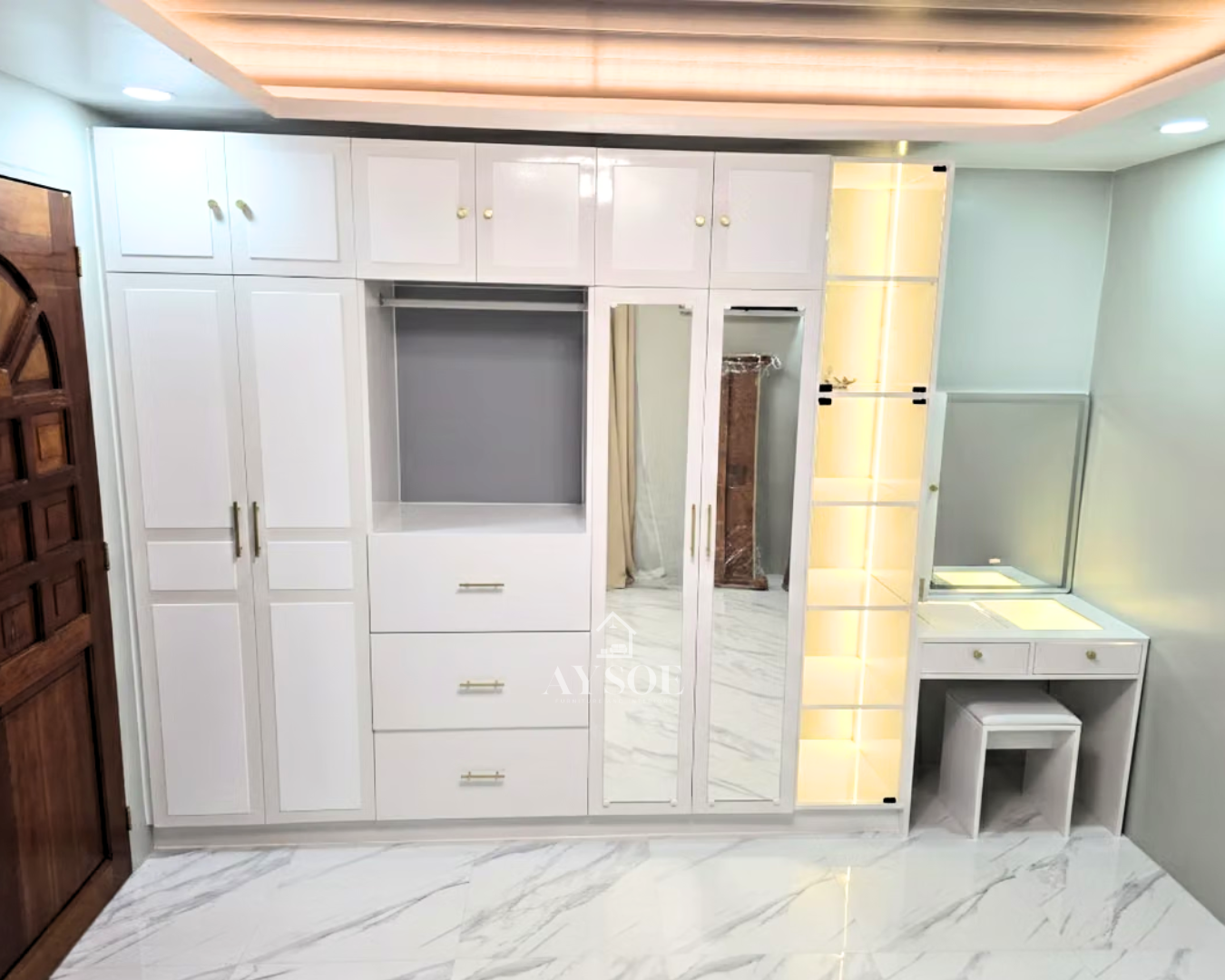 A floor-to-ceiling white shaker wardrobe with gold hardware, mirror doors, and drawers. A lit glass display tower and built-in vanity complete the wall.