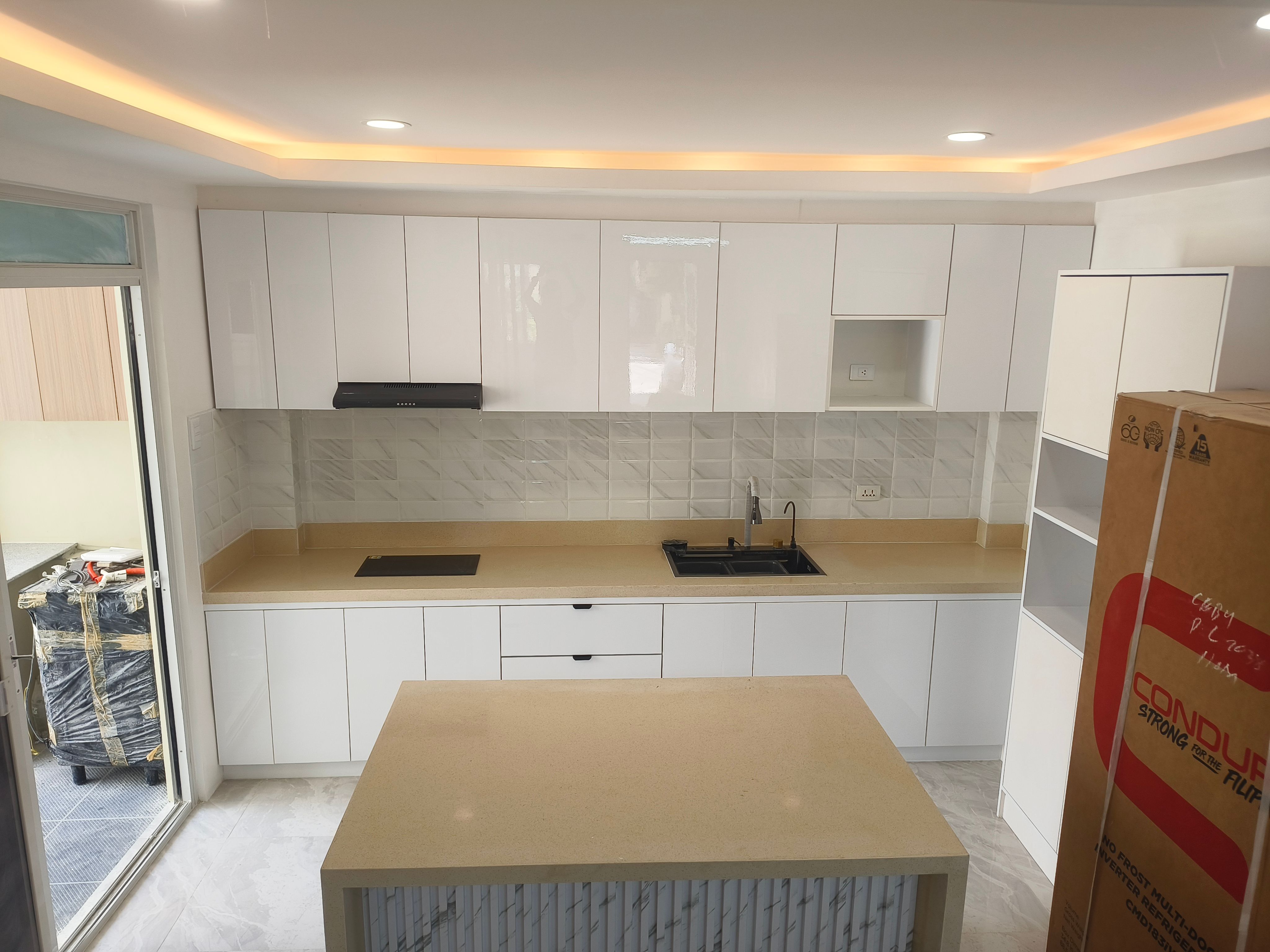 A full-room kitchen in high-gloss white with a waterfall island, sand-toned solid-surface counters, and a cove-lit tray ceiling. Floor-to-ceiling cabinetry, a built-in induction hob, and a tall pantry unit keep everything organized in a bright, open layout.