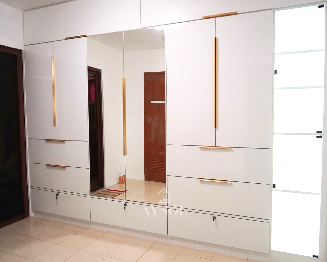 A floor-to-ceiling built-in wardrobe in high-gloss white, with gold bar handles, a full-height center mirror, roomy drawers, three locking drawers, and glass-door shelving. Made to fit your wall.