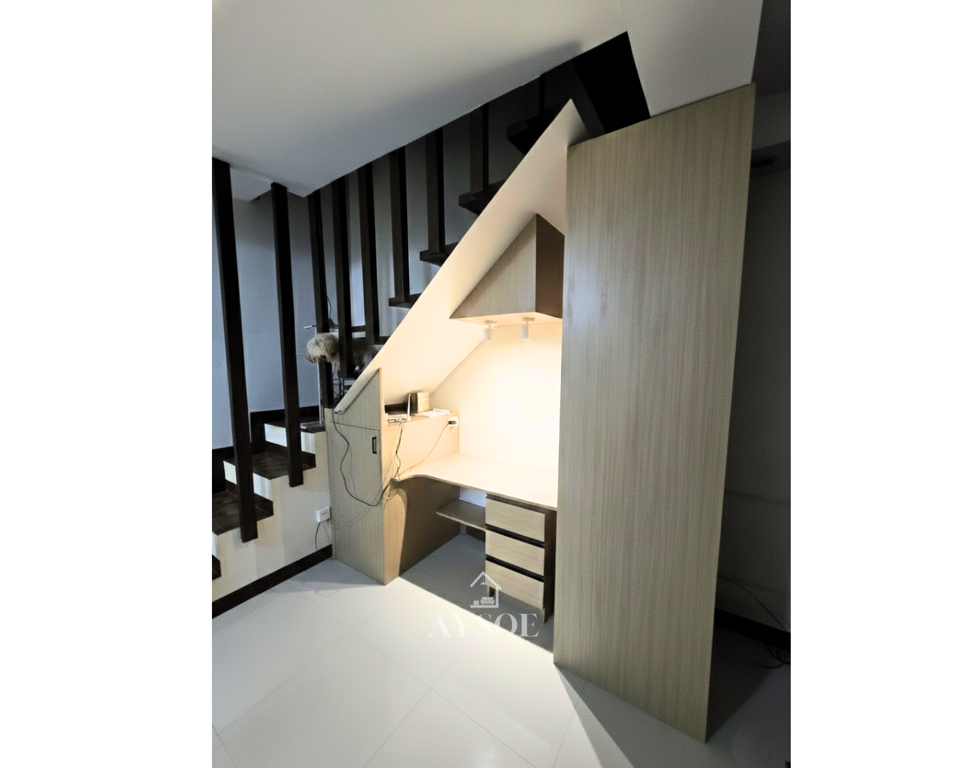 A custom under-stair desk nook in a light oak wood-grain finish, with a built-in desk, three drawers, a tall bookshelf, and a storage cabinet. Turns unused stair space into a home office.