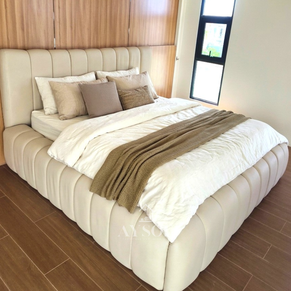 A soft, sculptural bed in smooth cream leatherette, with a tall wide channel headboard and thick pillow-style channels wrapping the whole frame. Plush, modern, and easy to wipe clean.