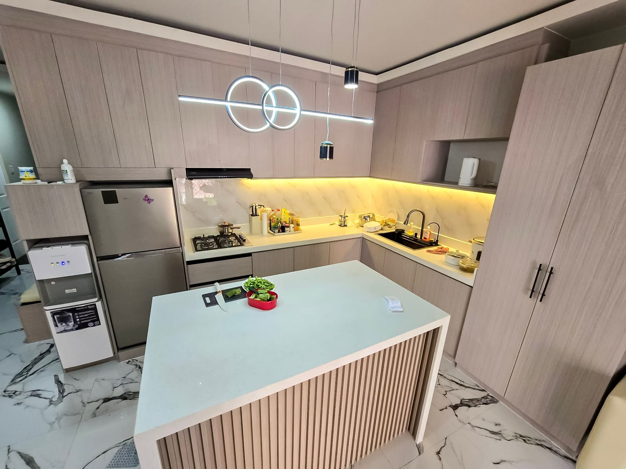 An L-shaped kitchen in soft taupe wood-grain cabinets, with a marble-look backsplash, warm LED lighting, and a white waterfall island with a wood-slat front. Tall storage and cabinets up to the ceiling make the most of the space.