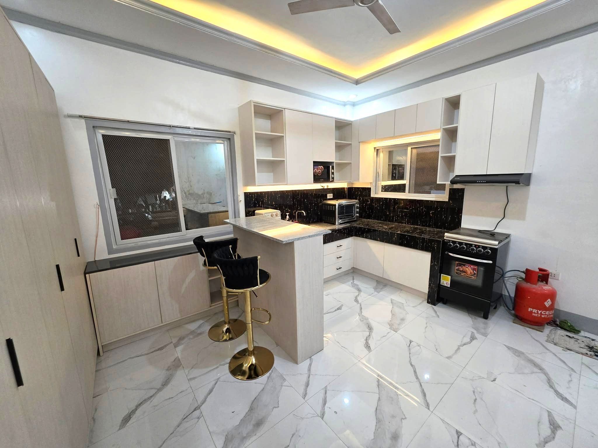 A cream wood-grain L-shaped kitchen with a black and gold backsplash, open shelves, and a granite-top breakfast bar.