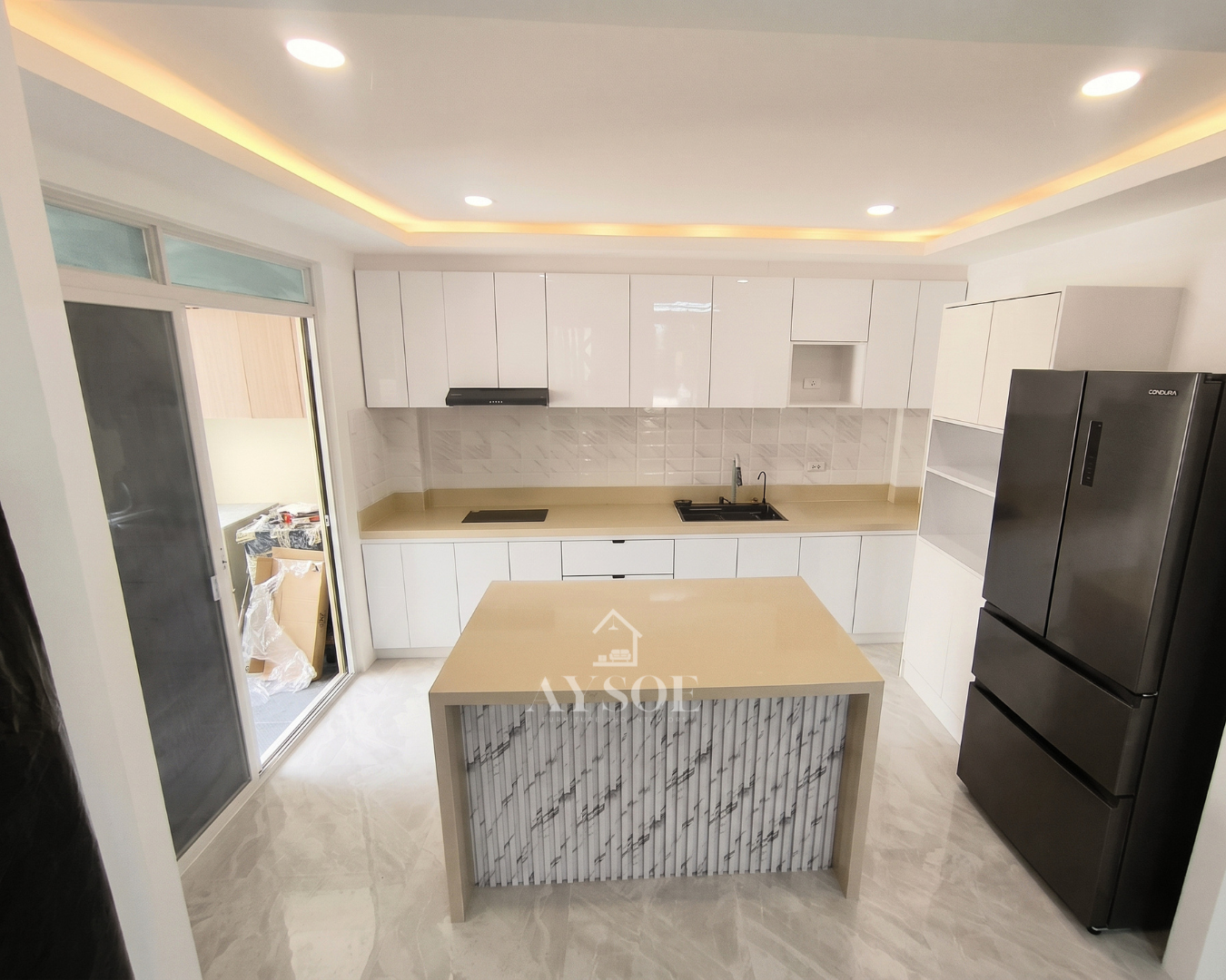 A high-gloss white kitchen with a waterfall island, sand-toned counters, and a cove-lit ceiling. Floor-to-ceiling cabinets, an induction hob, and a tall pantry keep it organized.
