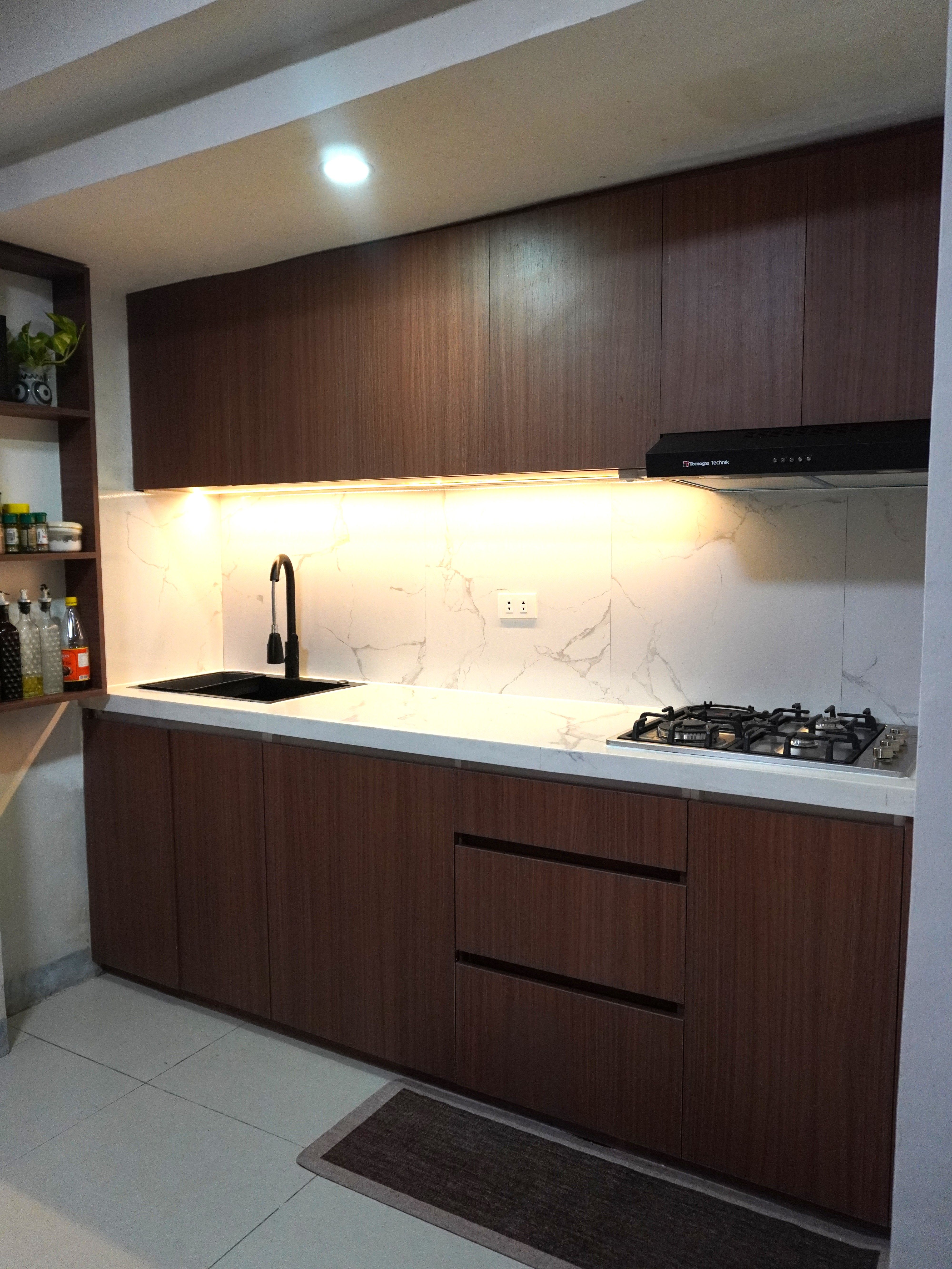 A warm single-wall kitchen in rich walnut-grain cabinetry with a Calacatta-style marble backsplash. Handleless doors, a black matte sink and faucet, and glowing under-cabinet lighting make it feel cozy and modern.