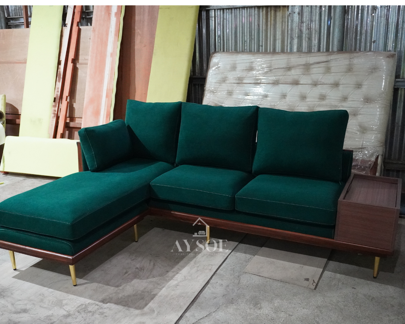 A mid-century L-shaped sofa in rich emerald velvet-style fabric, with a deep left chaise, plush loose cushions, a walnut base, brass-gold legs, and a built-in storage side table. Bold, comfortable, made to order.
