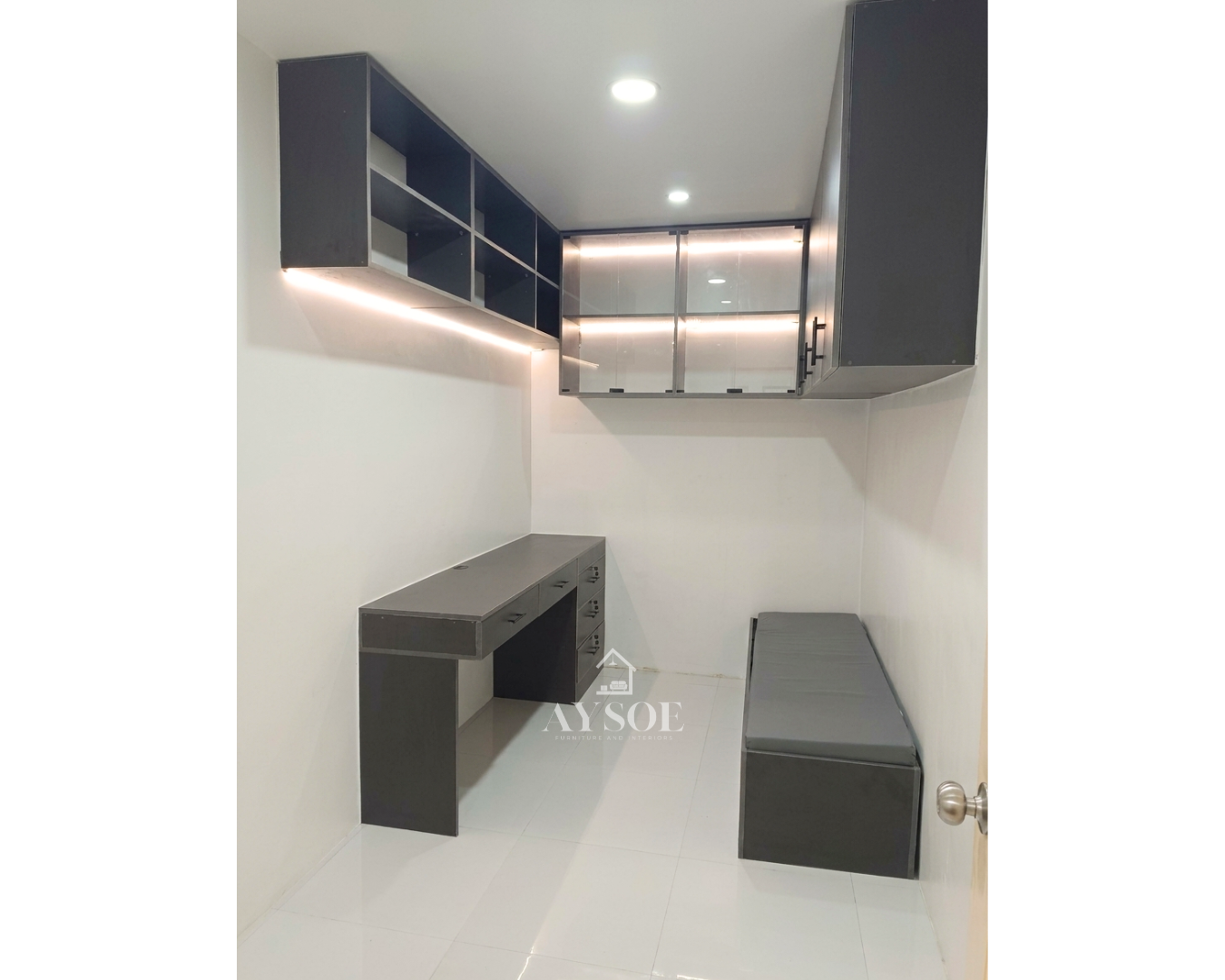A compact charcoal-grey home office set with a built-in desk, wall-mounted shelves, lighted glass display cabinets, and a cushioned bench. Great for small rooms, studies, and work-from-home spaces.