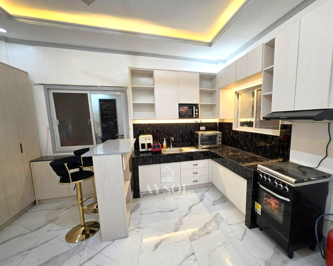 A cream wood-grain L-shaped kitchen with a black and gold backsplash, open shelves, and a granite-top breakfast bar.