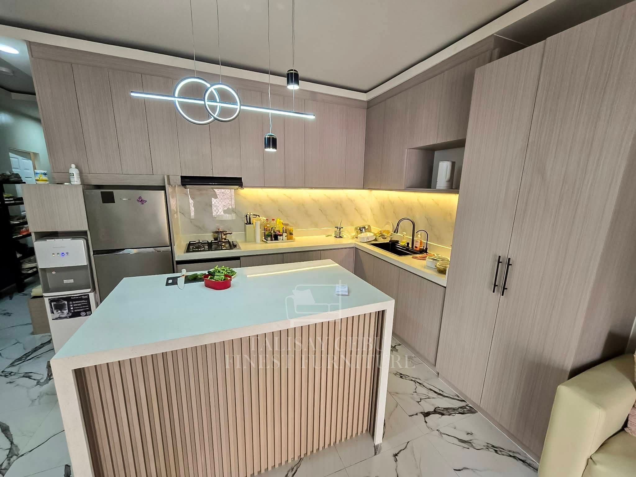 An L-shaped kitchen in soft taupe wood-grain cabinets, with a marble-look backsplash, warm LED lighting, and a white waterfall island with a wood-slat front. Tall storage and cabinets up to the ceiling make the most of the space.