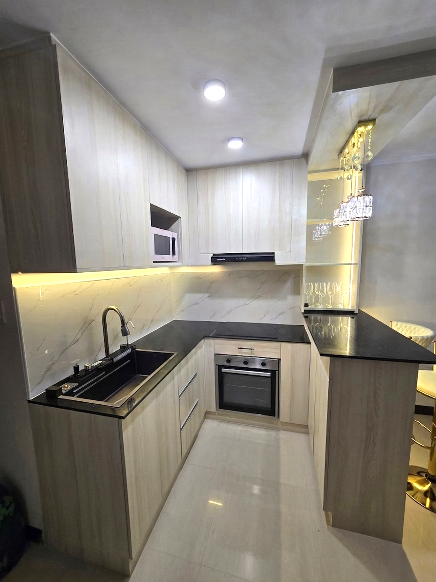 A compact light oak kitchen with a marble-look backsplash, breakfast bar, and lit glass display cabinet.