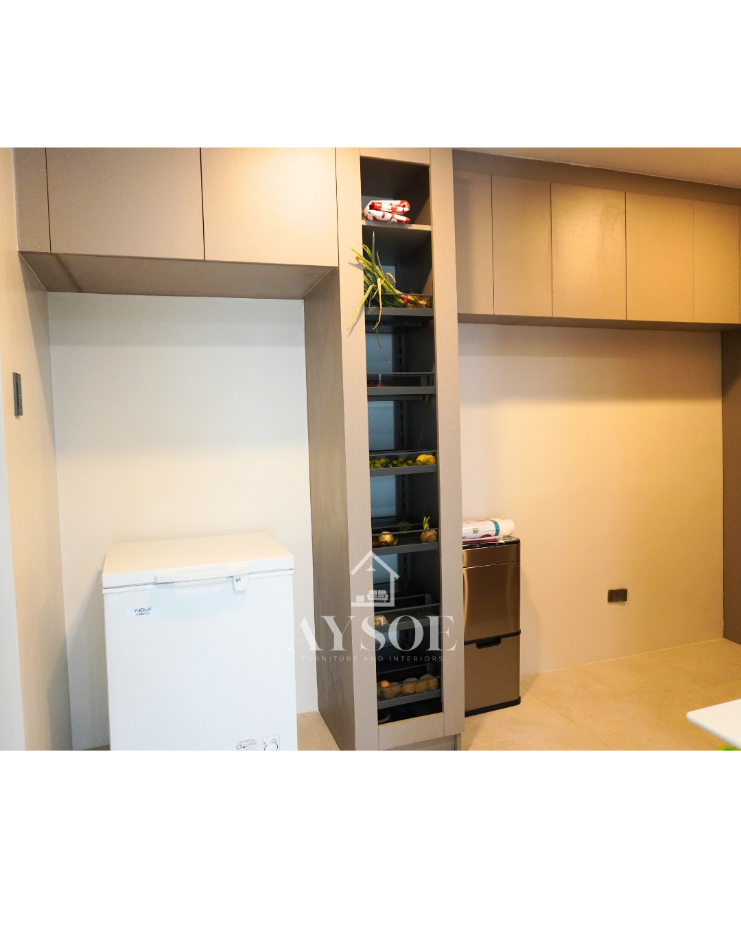 A custom taupe linen-finish storage set with a tall pull-out pantry with six wire baskets and handleless overhead cabinets. Organized storage for kitchens and utility areas.